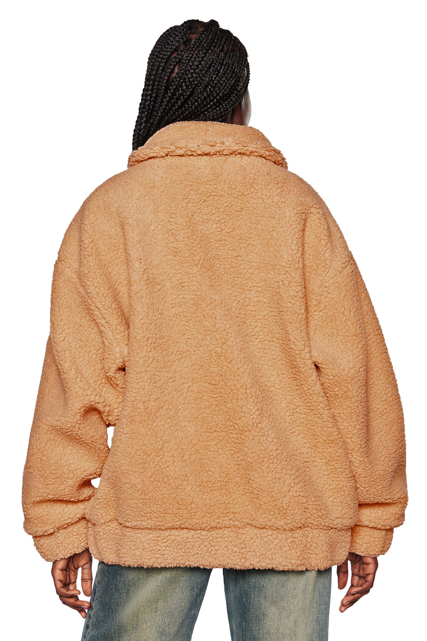 A person with braided hair is viewed from behind, donning the I.AM.GIA PIXIE COAT in caramel, an oversized plush fleece jacket. Their arms rest casually by their sides, paired elegantly with light blue jeans. The plain white background accentuates the casual yet stylish outfit.