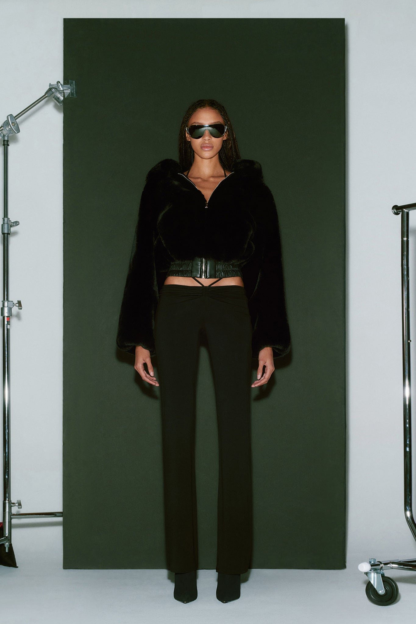 A person stands against a black background wearing the 717 JACKET - BLACK from I.AM.GIA, straight black pants, black shoes, and sunglasses. The outfit is accessorized with a belt. Studio lighting equipment is partially visible on the sides.