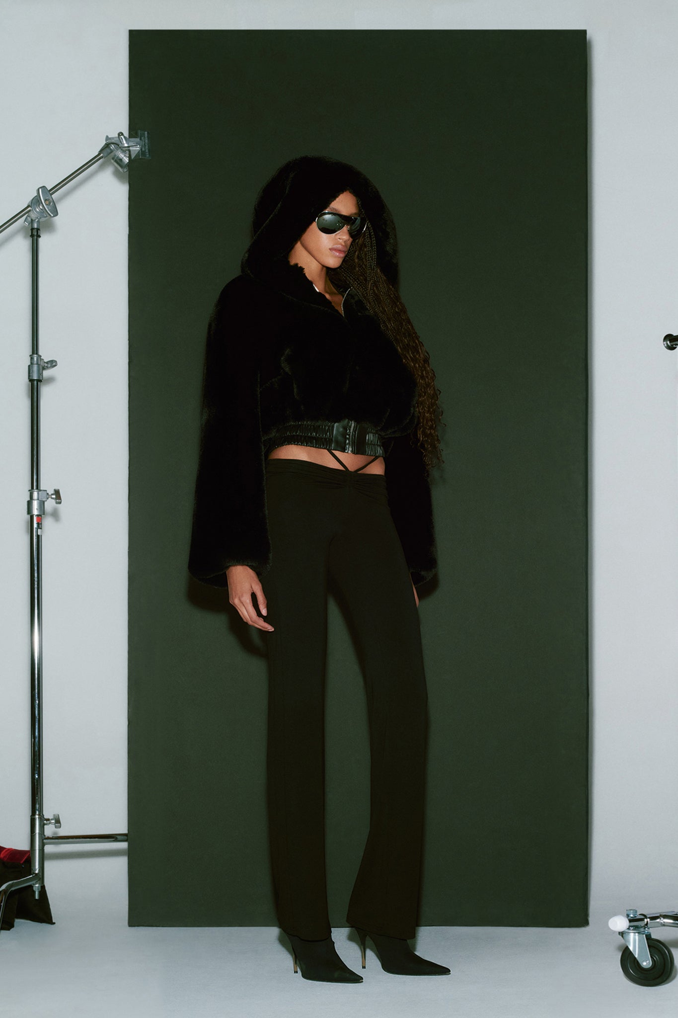 A model stands confidently in front of a green backdrop, wearing the I.AM.GIA 717 Jacket in black, paired with black sunglasses and black pants. They have long, curly hair and are completing their look with black pointed-toe heels. Studio lighting equipment is visible on the left side.