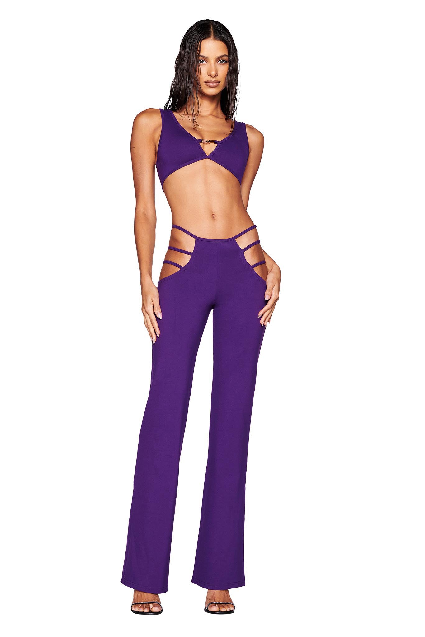 A person dressed stylishly in a purple two-piece ensemble by I.AM.GIA, reminiscent of a Euphoria Maddy outfit. It features a crop top paired with the LUCID PANT - PURPLE, showcasing cut-out designs on the hips. The look is finished off with black high heels.