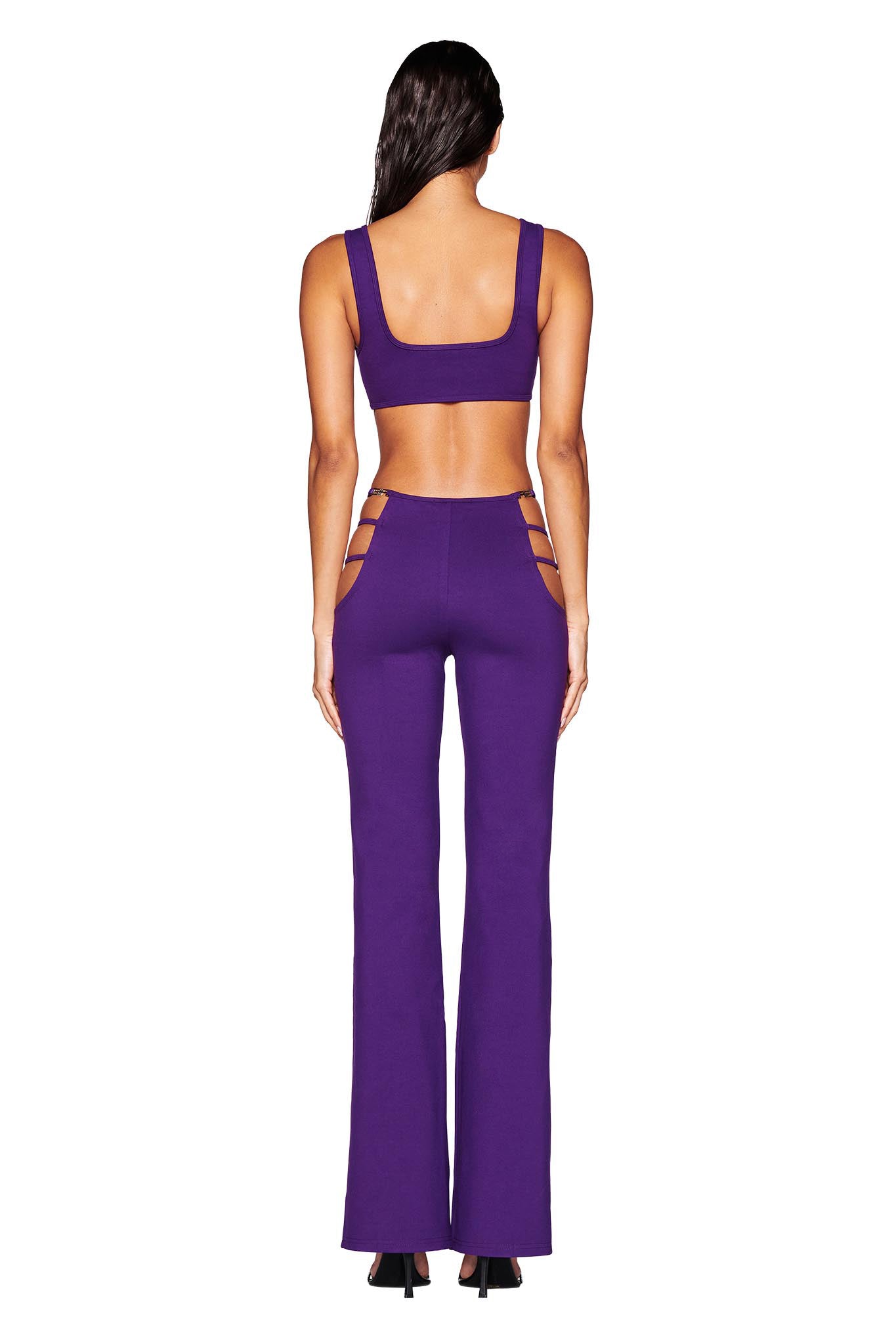 A person wearing the LUCID TOP - PURPLE from I.AM.GIA faces away, showcasing a sleeveless crop top paired with high-waisted flared pants that feature side cutouts. The ensemble is bodycon fit and is set against a plain white background.