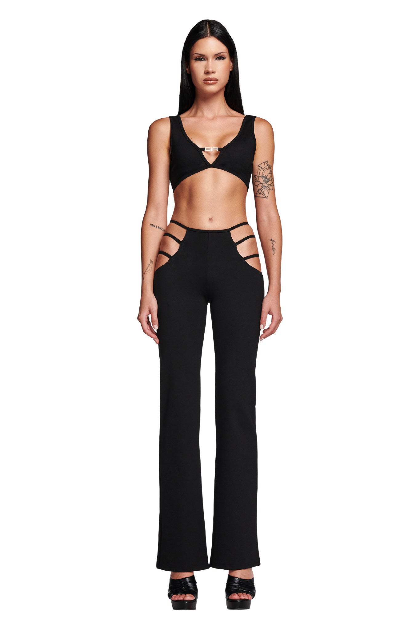 A person is wearing a black two-piece from I.AM.GIA, featuring a crop top and the high-rise LUCID PANT - BLACK, which showcases side hip cut-outs. They are standing against a white background, with their long hair and tattoos visible on one arm.