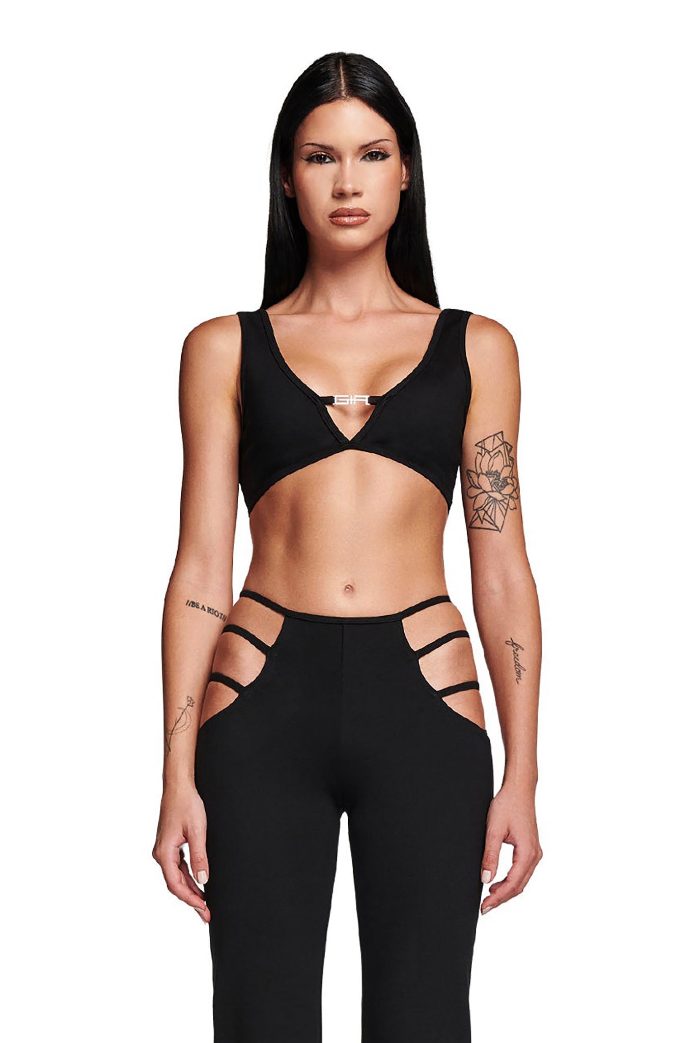 A person with long dark hair is seen wearing the black LUCID TOP by I.AM.GIA and high-waisted pants featuring cut-out designs, which reveal their arm tattoos. The bodycon fit enhances their edgy style against a plain white background.
