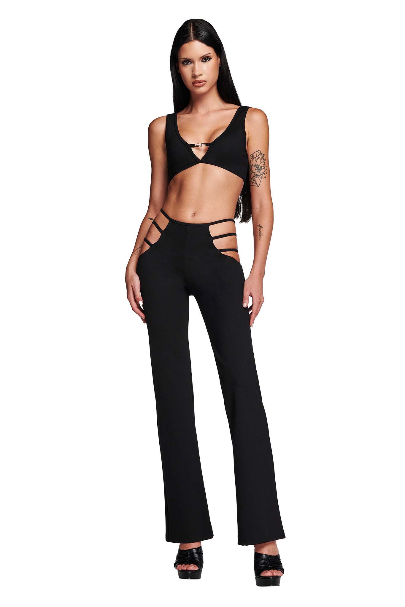 A woman in a black cut-out top and high-rise I.AM.GIA LUCID PANT - BLACK stands confidently. Her long hair pairs well with her stylish high-heeled sandals. The pants, featuring side hip cut-outs, add a chic touch, while a tattoo graces her upper arm against the white background.