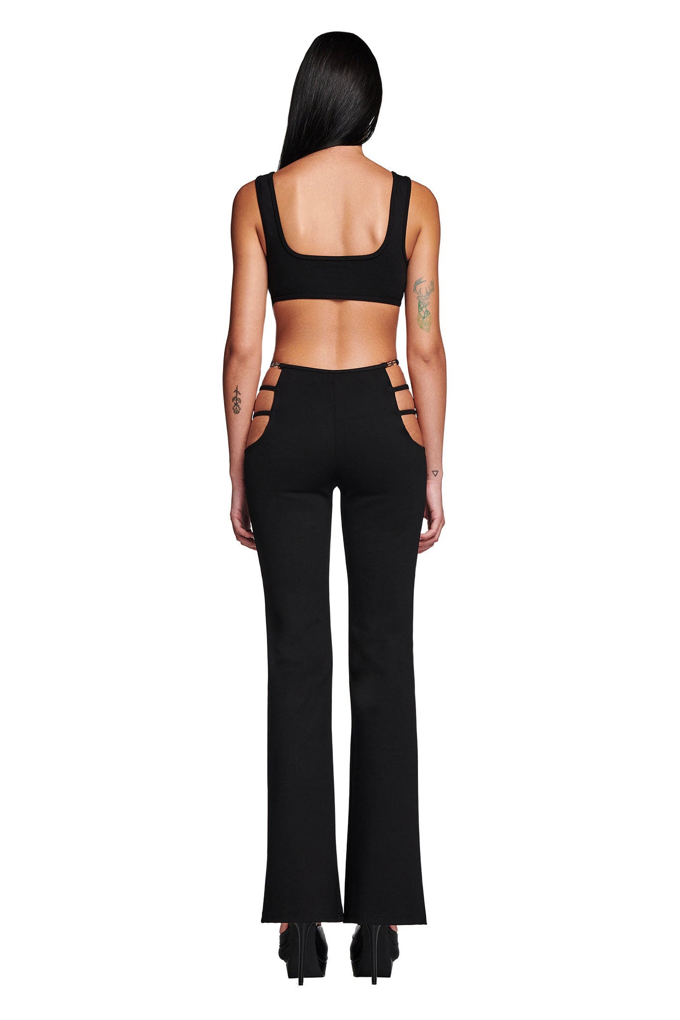 A woman with long black hair is wearing a sleek LUCID TOP in black by I.AM.GIA, paired with high-waisted pants featuring cut-out details on the sides. The bodycon fit highlights her silhouette as she poses with her back to the camera against a plain white background.