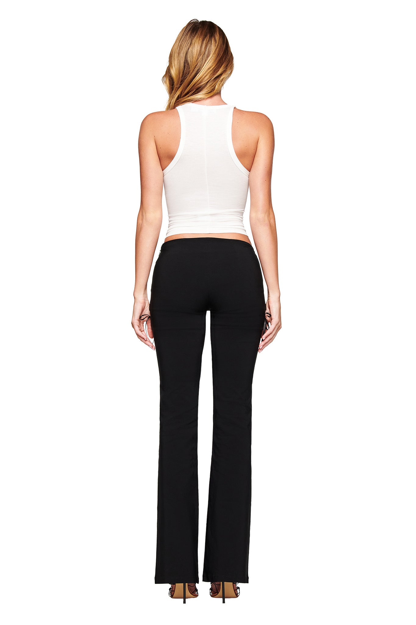 A blonde woman stands facing away on a white background, wearing the I.AM.GIA PENELOPE TANK - PSYCHO, a white ribbed tank top, paired with black flared pants.