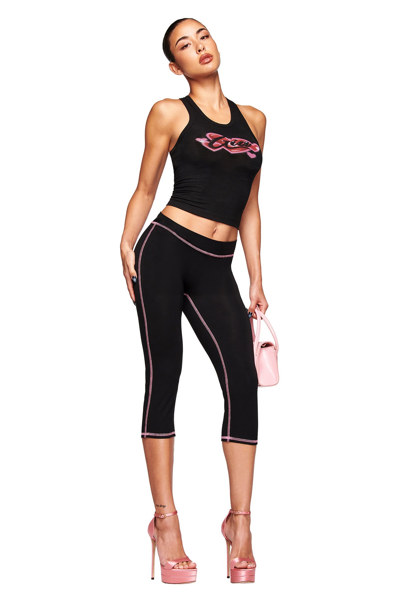 A woman poses in the I.AM.GIA PENELOPE TANK - GIA AIRBRUSH with a pink graphic, black capri leggings with pink piping, pink high heels, and a small pink handbag, standing against a white background.