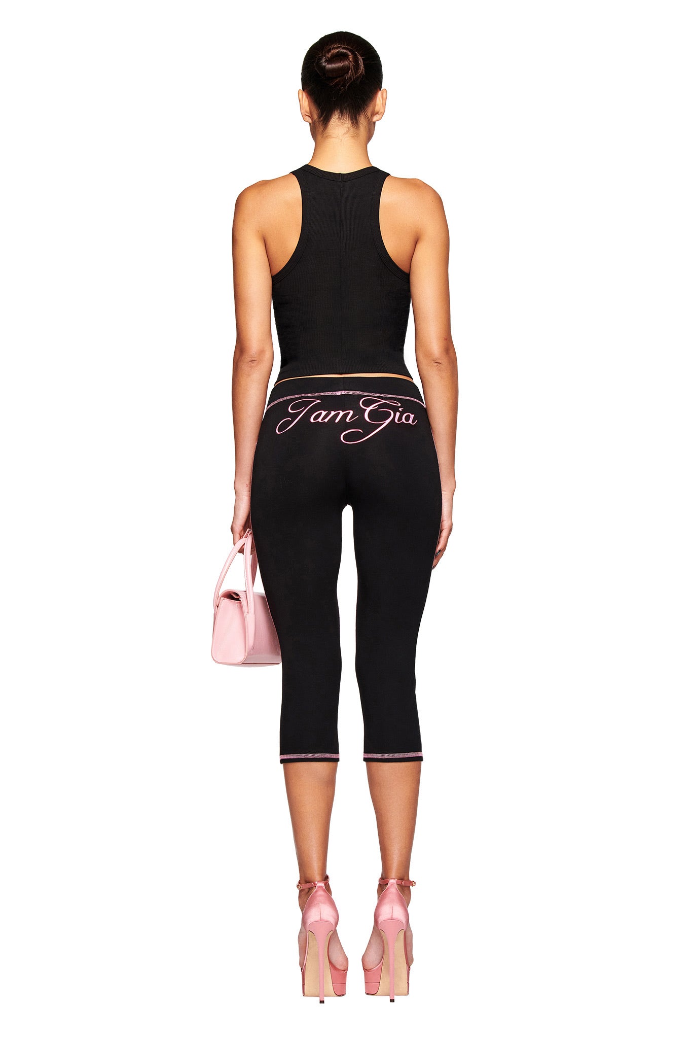 A woman faces away, wearing the black PENELOPE TANK - GIA AIRBRUSH by I.AM.GIA and black cropped leggings with pink "I Am Gia" on the back. She carries a pink handbag, wears pink heels, and her hair is in a bun.