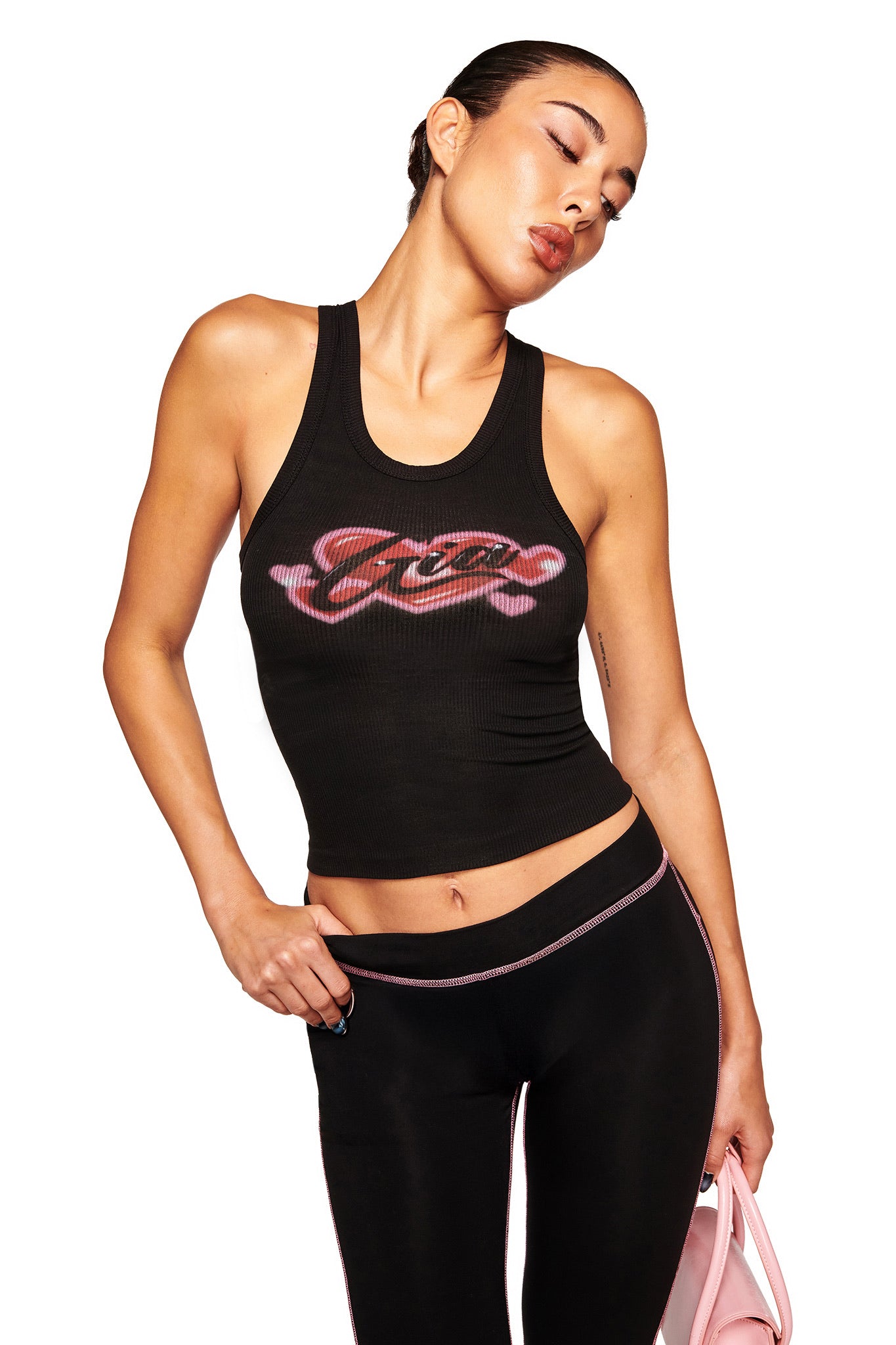 A woman with slicked-back hair poses serenely, eyes closed, wearing the I.AM.GIA PENELOPE TANK - GIA AIRBRUSH featuring a lips and bone graphic, paired with black leggings with pink trim, and holding a pink bag.