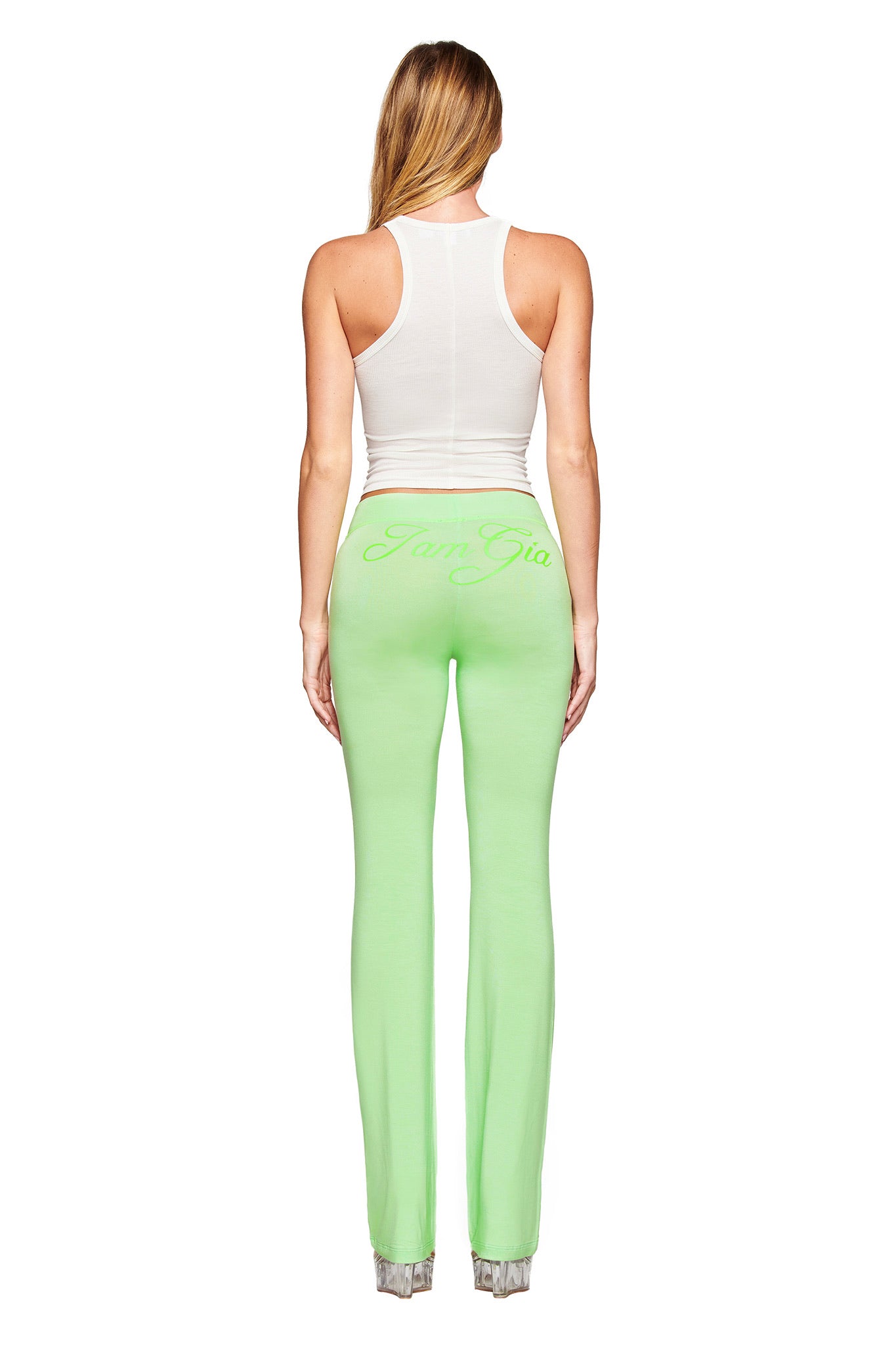 A woman faces away, wearing the I.AM.GIA PENELOPE TANK - STAR AIRBRUSH and light green fitted pants with "IamGia" on the back. The bodycon fit and plain white background accentuate her modern style.