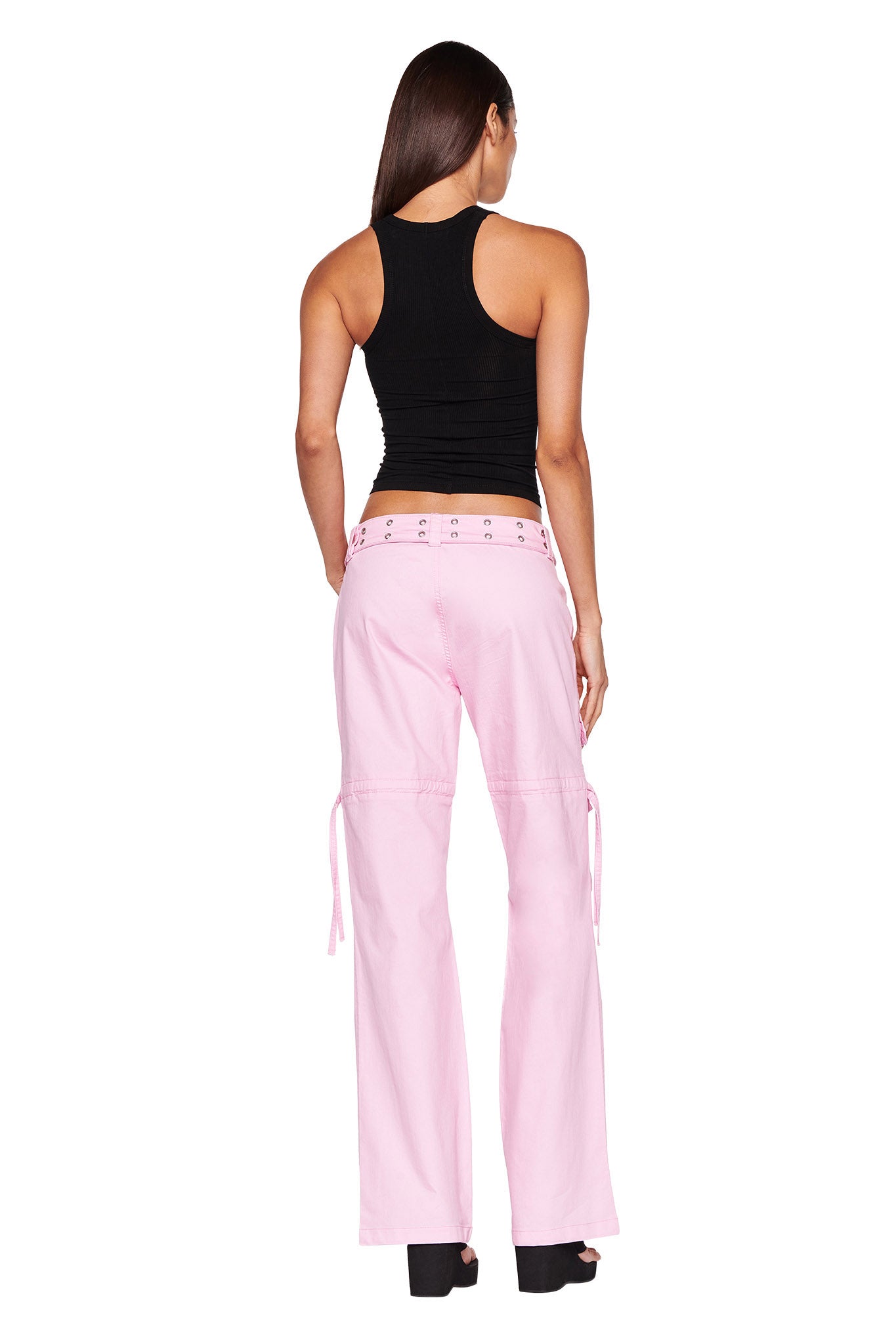 A woman with long brown hair stands with her back to the camera, showcasing a black sleeveless top and light pink NAIMA CARGO PANTS - PINK BABY from I.AM.GIA that have tie details on the sides. She completes the look with black platform shoes. The background is completely white.