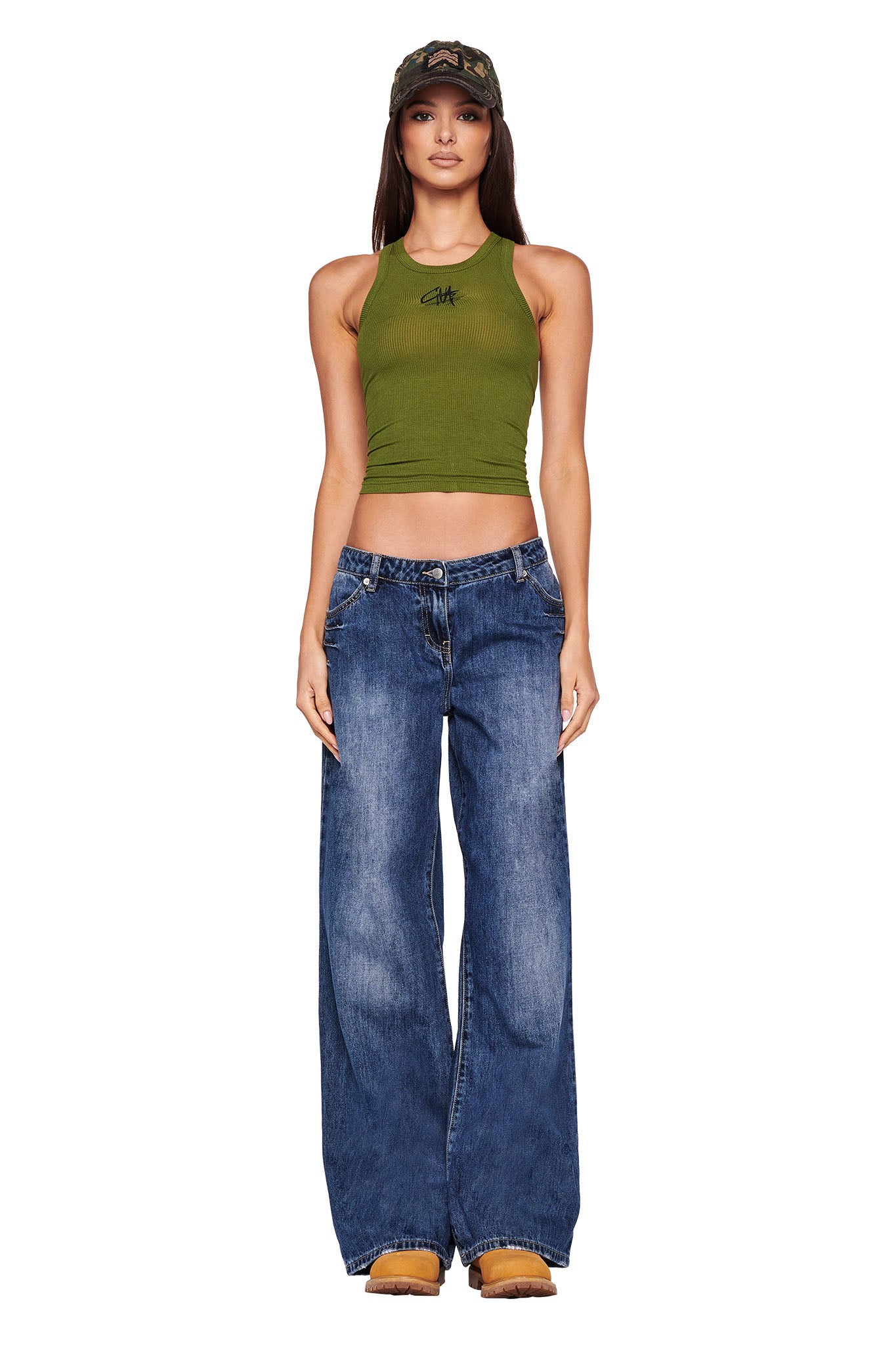 A woman stands against a white background, wearing an I.AM.GIA green cropped halter top with a small black graphic, wide-leg DYLAN JEAN - BLUE : DARK BLUE from I.AM.GIA, and tan boots. She has long dark hair and wears a camouflage cap.