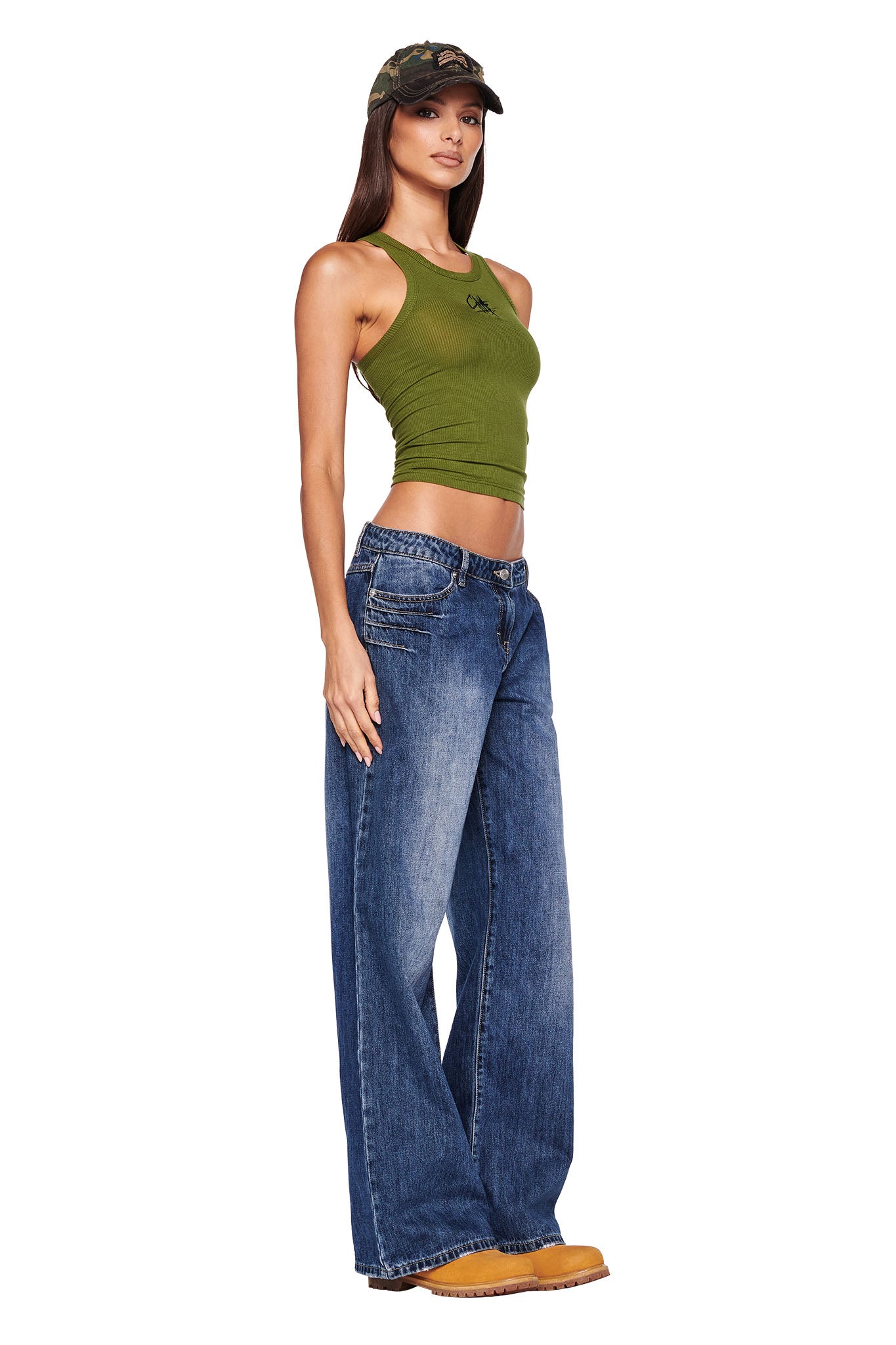 A woman stands wearing a green tank top, I.AM.GIA's DYLAN JEAN in dark blue, yellow boots, and a camouflage cap. She faces forward at a slight angle with one hand resting on her thigh against a plain white background.