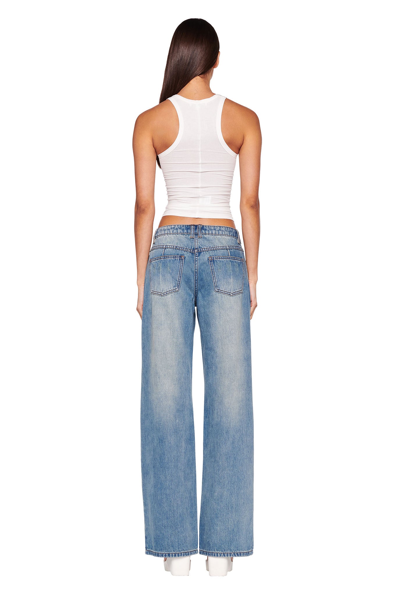 A woman with long, straight hair is standing with her back to the camera, showcasing her I.AM.GIA white sleeveless tank top and DYLAN JEAN - BLUE light blue wide-leg jeans from I.AM.GIA. She completes the look with stylish white shoes.