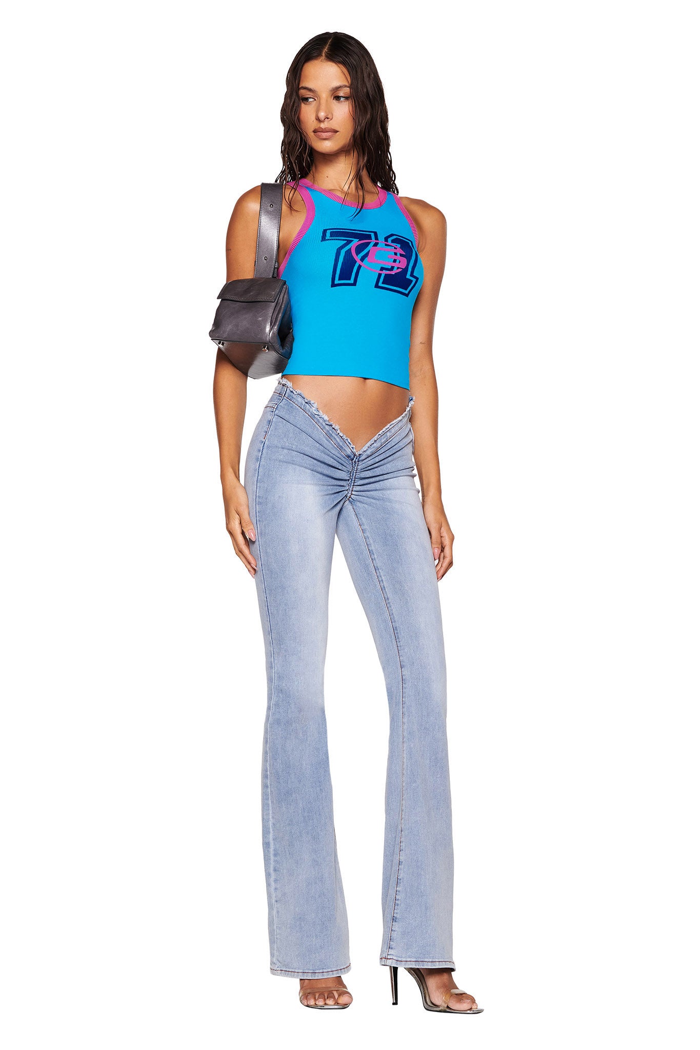 A woman poses confidently in front of a white backdrop, wearing the PENELOPE TANK - BLUE by I.AM.GIA, paired with light blue jeans that have a V-shaped waistband and high heels. She carries a black shoulder bag, her long wavy hair adding to her effortlessly stylish look with the soft ribbed fabric outfit.