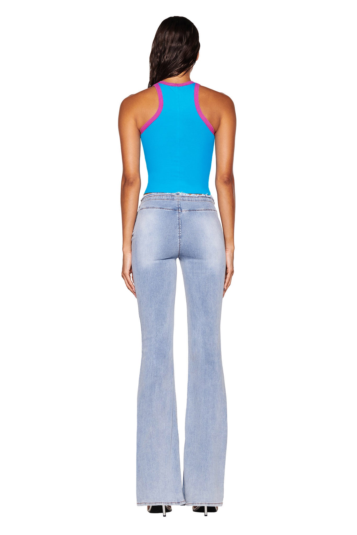 A person wearing an I.AM.GIA Penelope Tank in sky blue paired with pink accents and light blue flared jeans stands facing away. The racer's tank style accentuates their long dark hair, beautifully complemented by black shoes against a white background.
