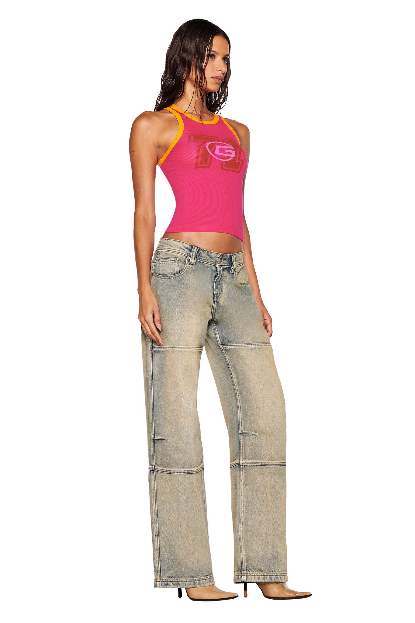 A person with wet, wavy hair is wearing a hot pink Penelope Tank by I.AM.GIA that showcases a custom graphic. They have paired it with light blue wide-leg jeans and brown high-heeled shoes, posing sideways with a slight turn towards the front.