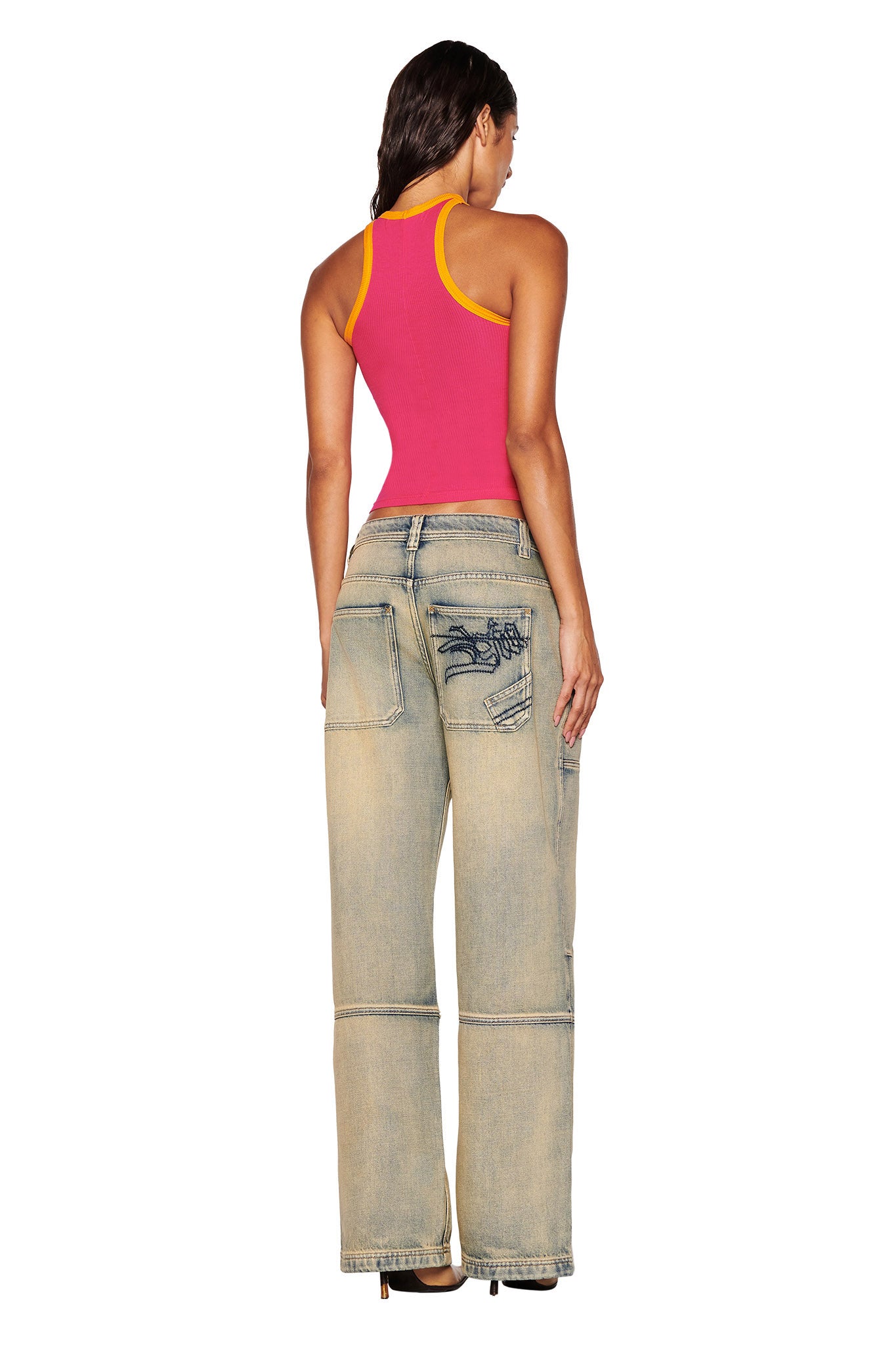 A person with long dark hair stands facing away, wearing the I.AM.GIA PENELOPE TANK in hot pink—a pink racer tank with yellow trim—and wide-leg, light blue jeans. The jeans feature unique stitching and custom graphic patchwork designs on the back.