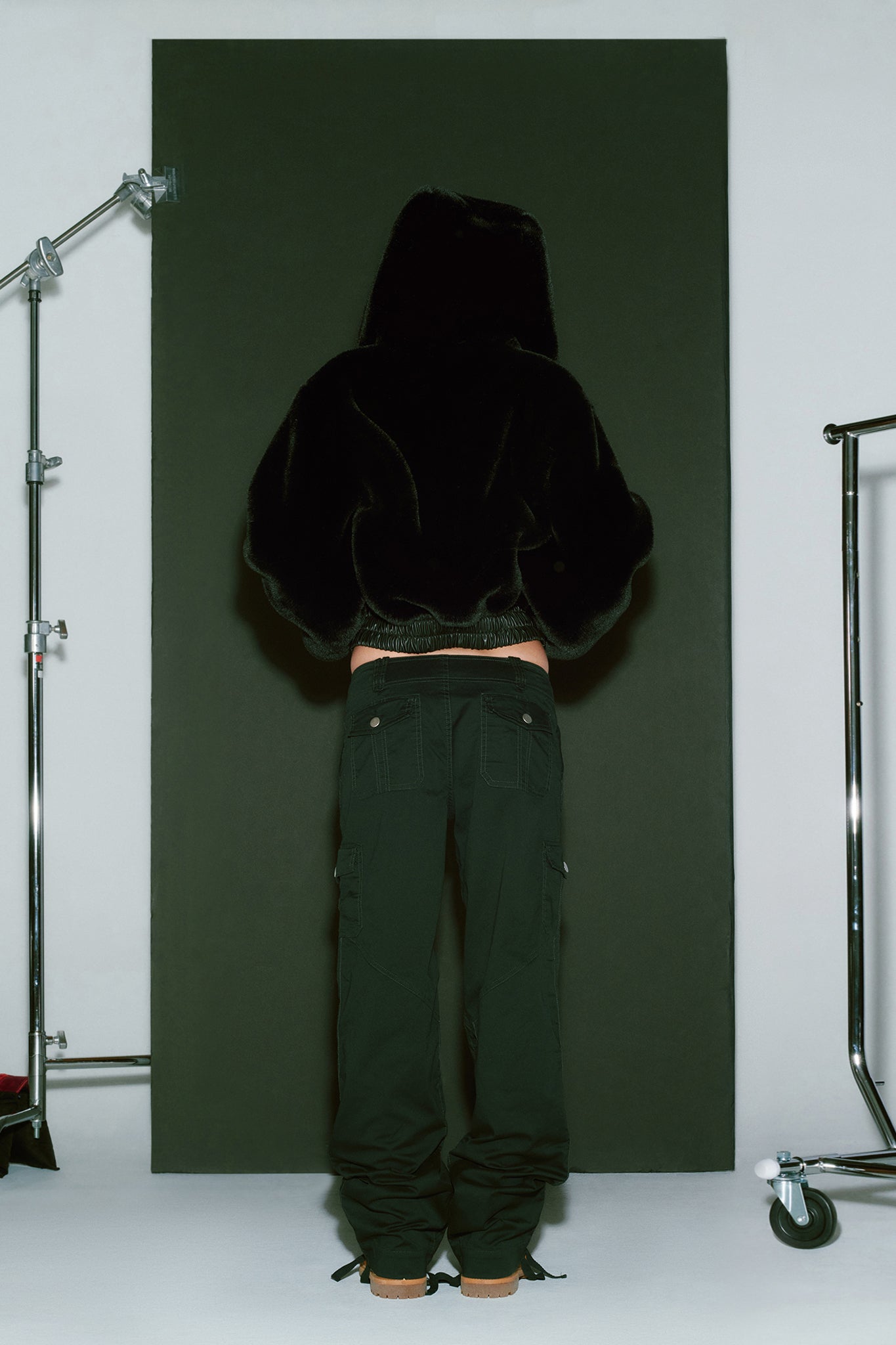 A person stands with their back to the camera, facing a black backdrop. They are wearing the I.AM.GIA 717 JACKET - BLACK and dark cargo pants with brown shoes. The surrounding area includes metal stands and lighting equipment, indicating a studio setup.
