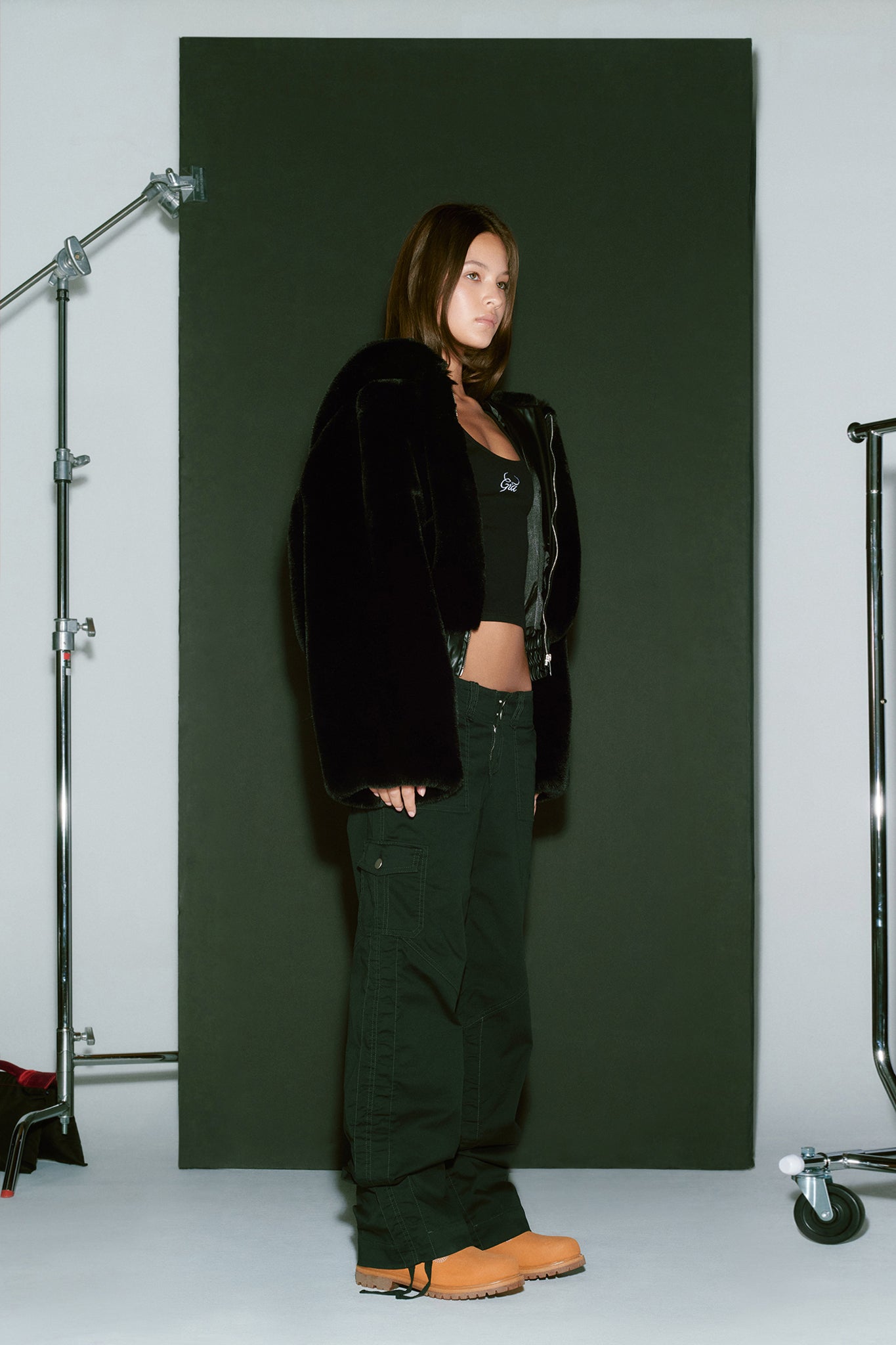 A person with long hair stands in front of a dark backdrop wearing the black 717 Jacket by I.AM.GIA, alongside a black top, black cargo pants, and light brown boots. They are posed with their hands by their sides and are surrounded by metal stands and equipment.