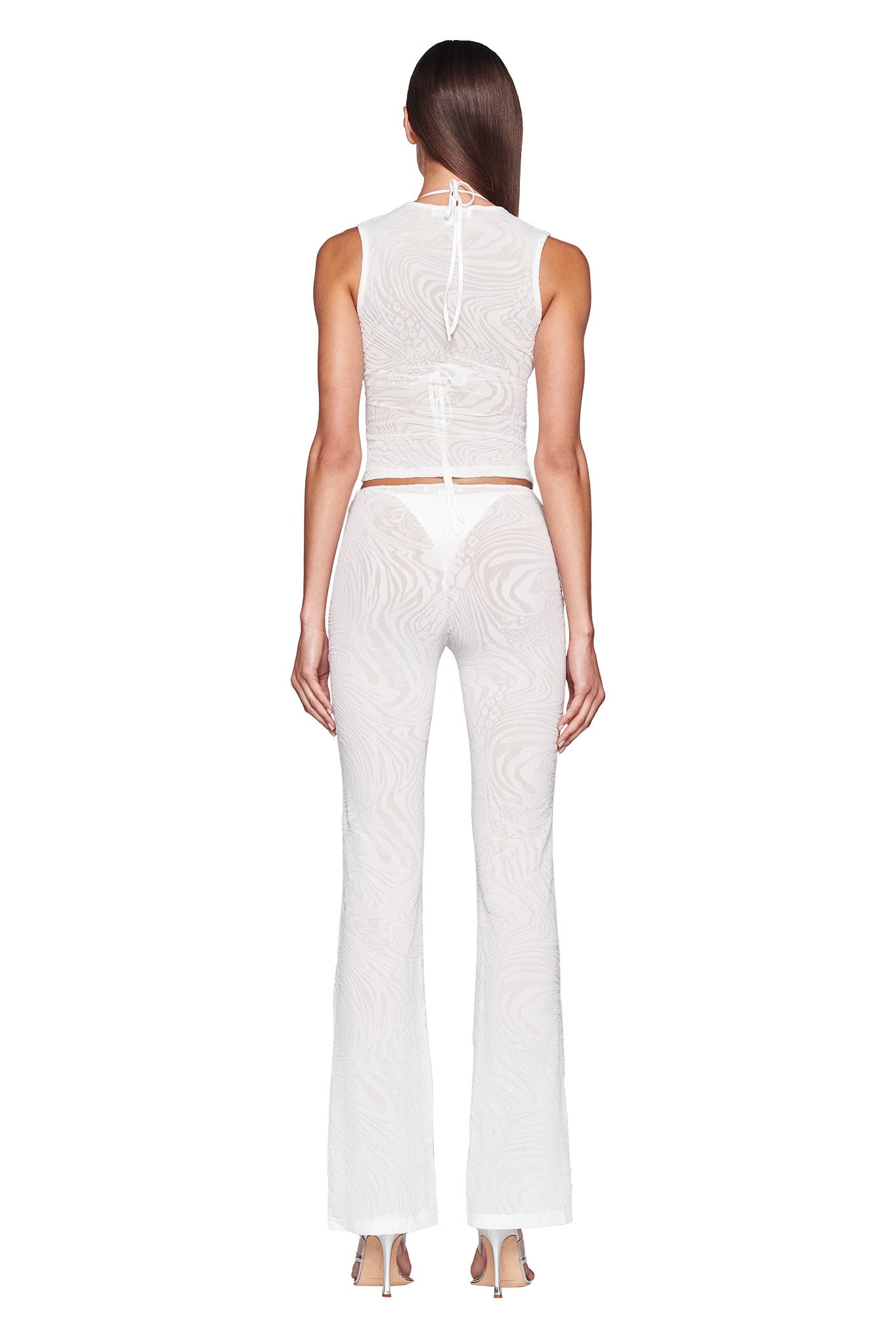 A model is standing with her back to the camera, wearing the I.AM.GIA AMINA PANT in white along with a matching white burnout velvet print sheer top. The outfit reveals white undergarments underneath. She has long, straight hair and is wearing high heels against a plain white background.