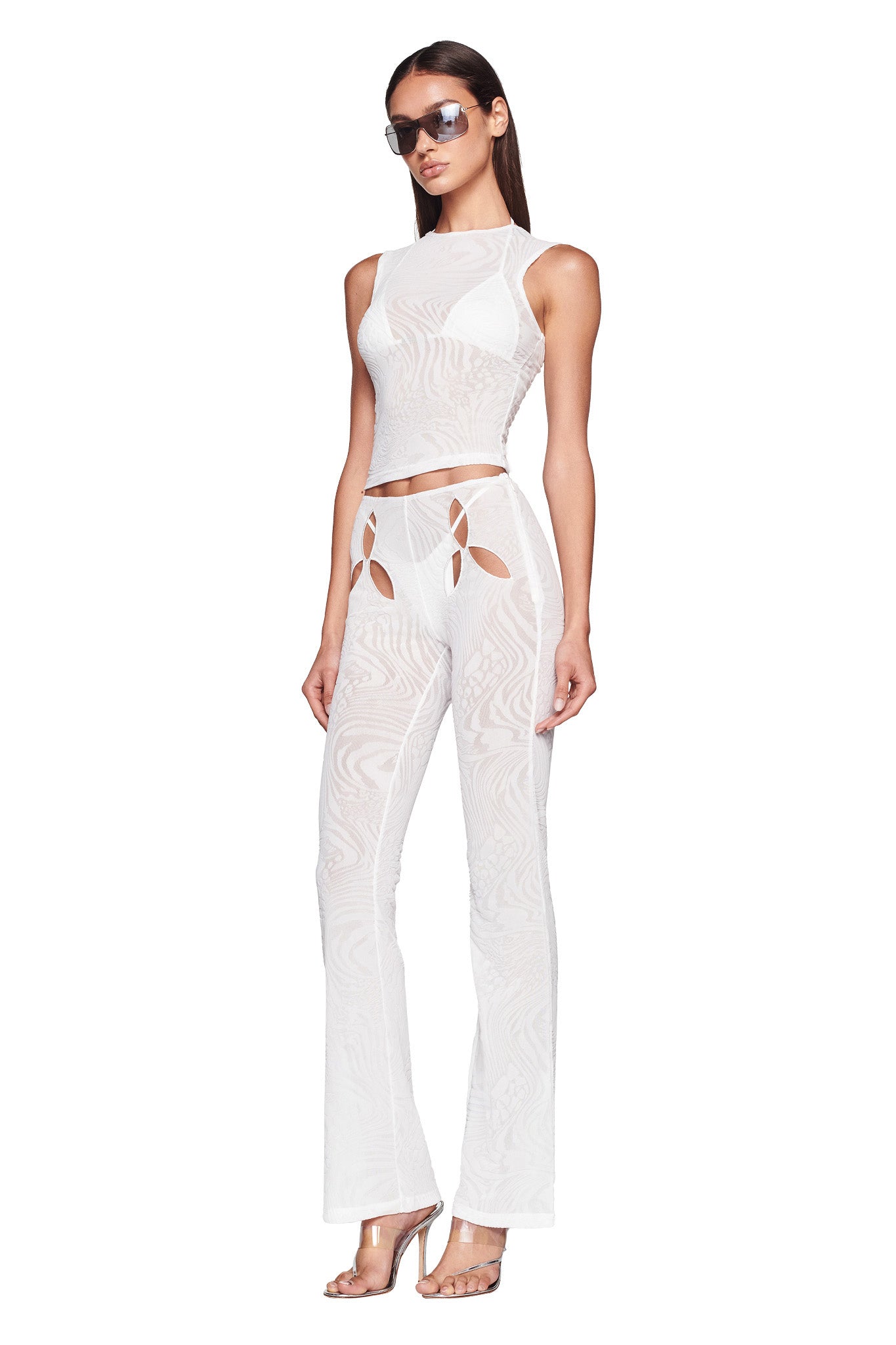 A woman with long hair wears sunglasses, a sleeveless sheer white top, and the AMINA PANT - WHITE from I.AM.GIA, featuring cut-out designs around the hips. She also wears high-heeled sandals and poses with a neutral expression against a plain white backdrop.