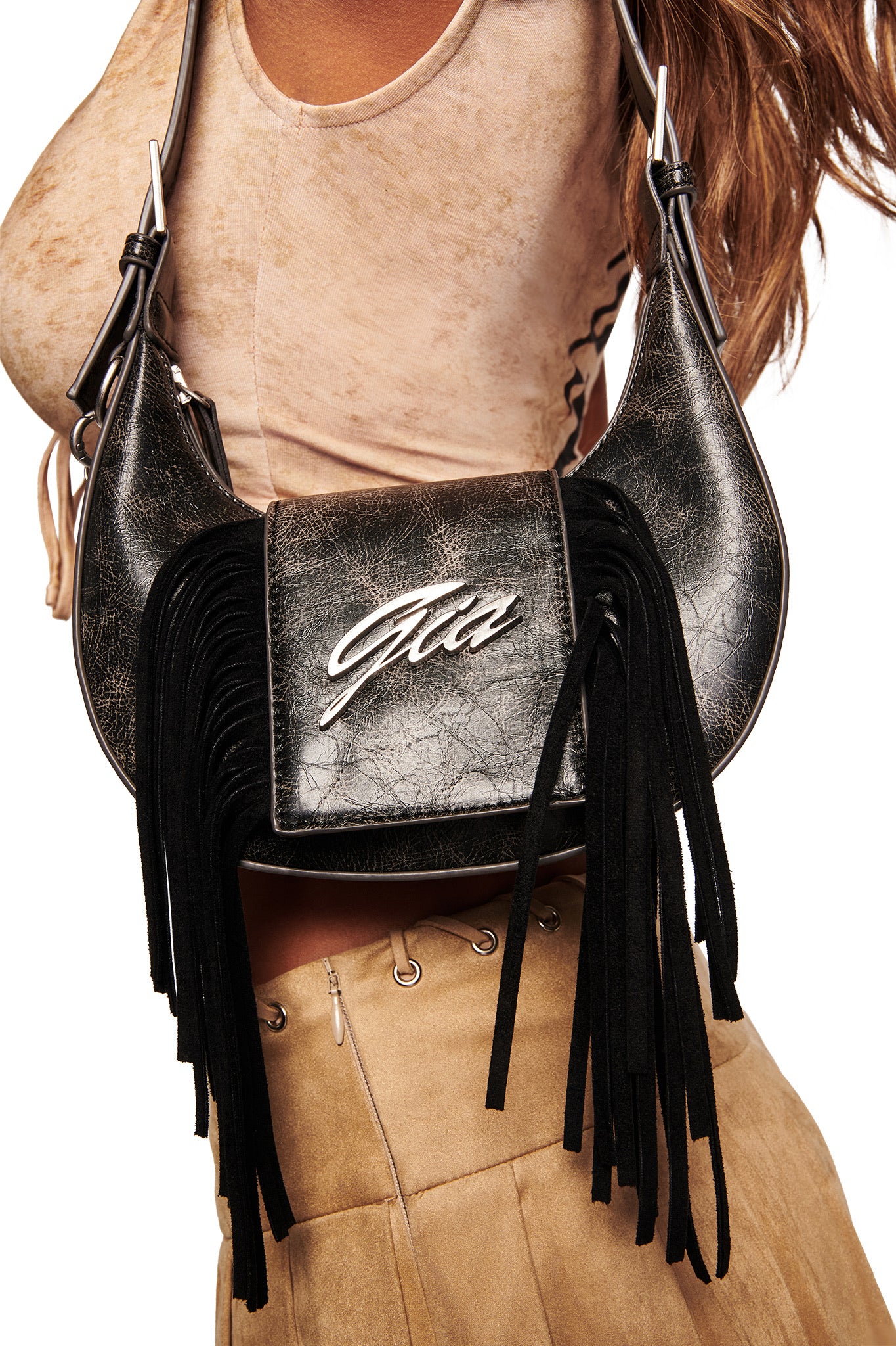 Close-up of a person wearing the I.AM.GIA TIA BAG - BLACK FRINGE, featuring fringe detail, a metallic logo, and an adjustable shoulder strap, paired with a beige top and tan suede skirt. White background.