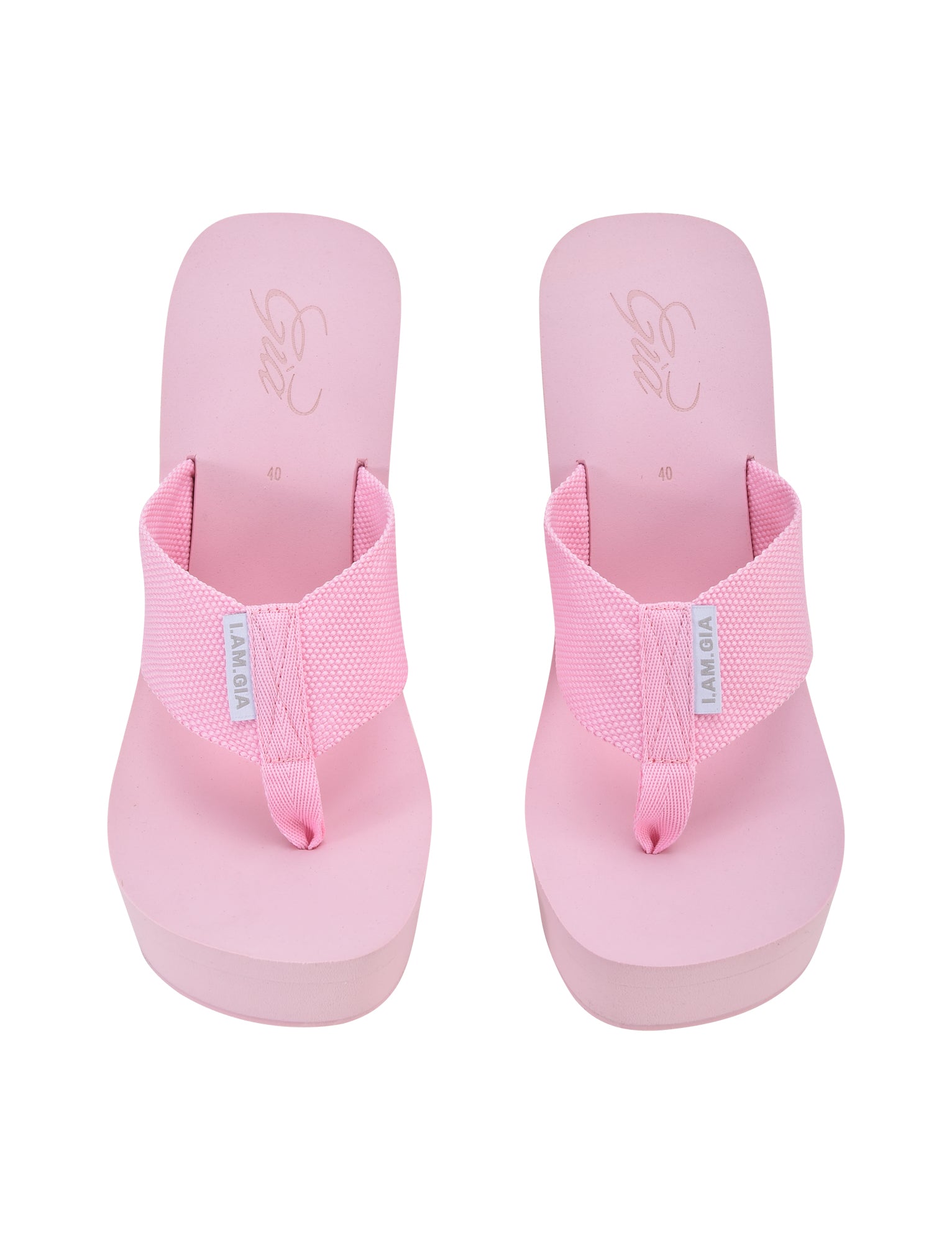 A pair of SHELBY FLIP FLOPS in baby pink from I.AM.GIA featuring thick straps and a toe thong design. The straps have a small white tag with the I.AM.GIA text, and the footbeds display cursive print near the heel area. The image is captured from a top-down view.