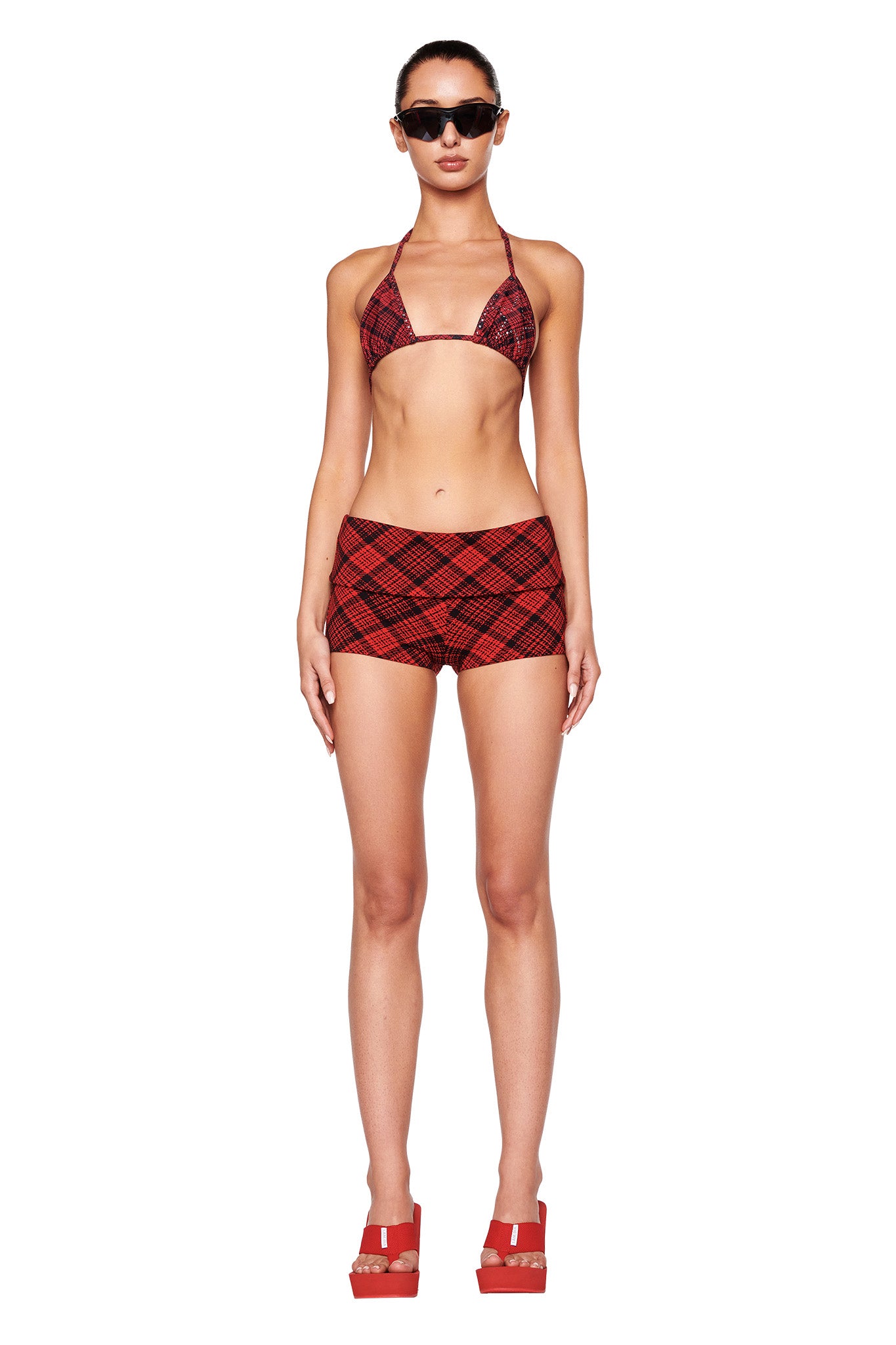 A person stands facing the camera wearing an I.AM.GIA red and black checkered bikini top, high-waisted shorts, and SHELBY FLIP FLOP - RED. They have short hair tied back, wear black sunglasses, and maintain a neutral expression. The background is plain white.