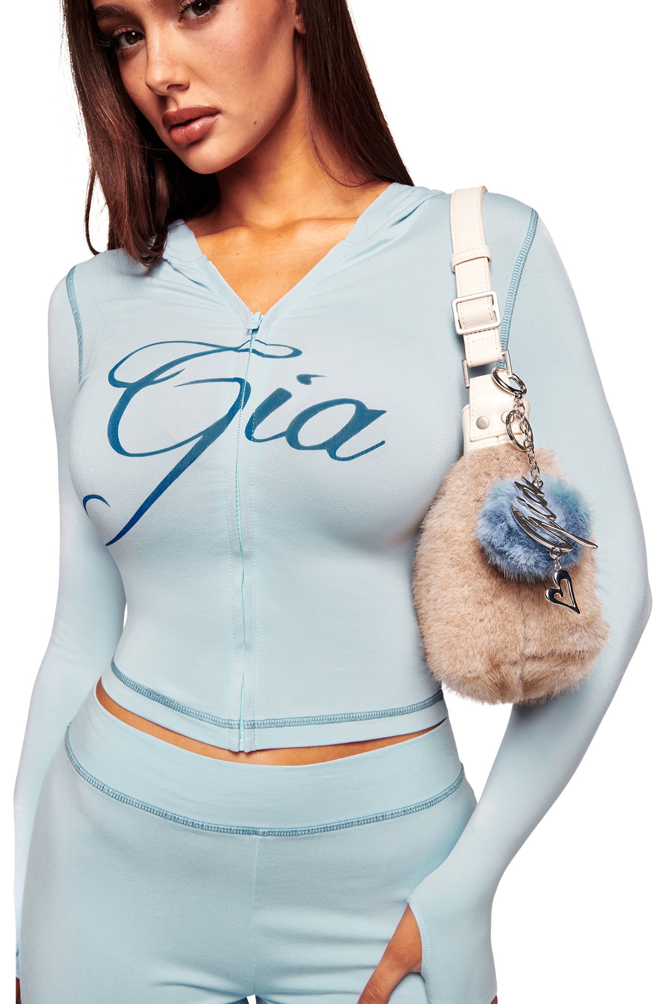 A woman wears a light blue fitted I.AM.GIA outfit with the GIA logo on her top. She carries a beige and blue furry bag featuring the LARSA KEYCHAIN - BLUE, standing against a white background.
