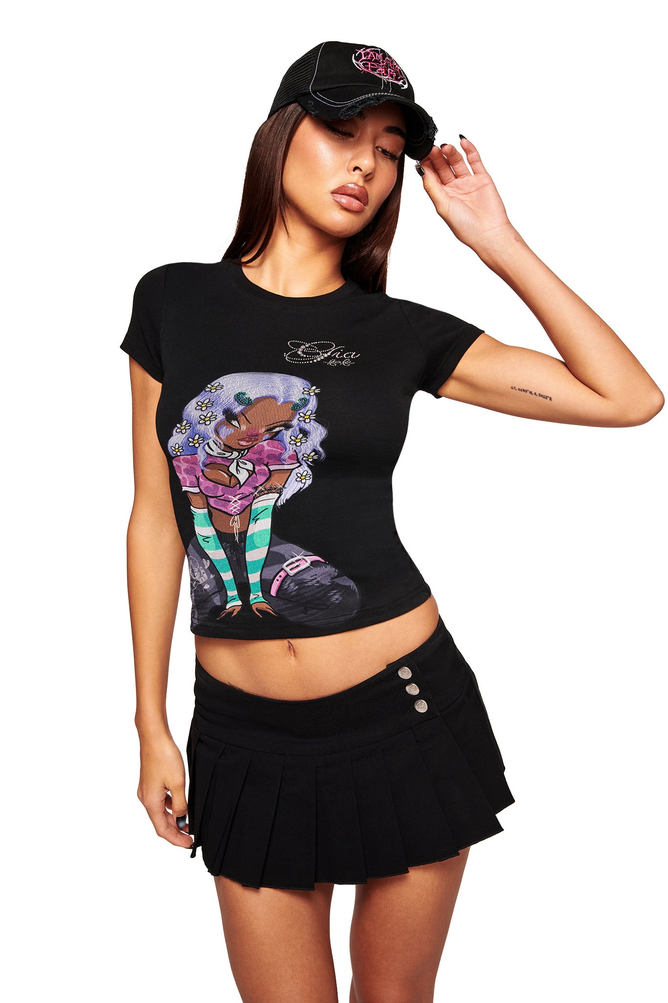 A woman poses against a white background, wearing the I.AM.GIA FAFI HAT - BLACK, an I.AM.GIA x FAFI black graphic tee with a colorful illustration, and a black pleated mini skirt, touching her cap with one hand.