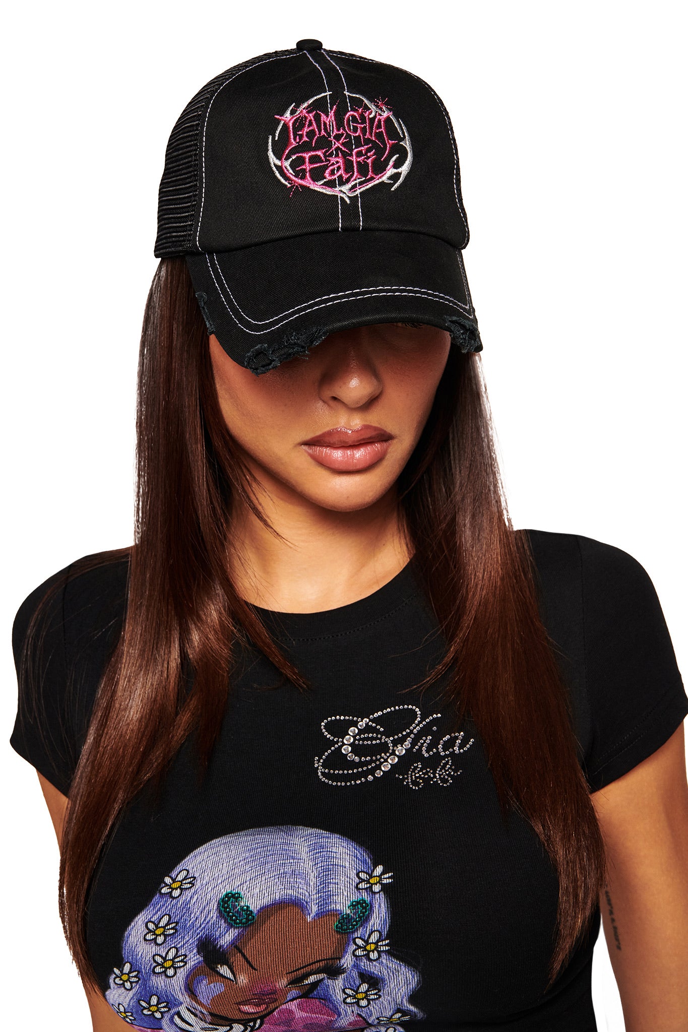A woman with straight, long brown hair wears the FAFI HAT - BLACK by I.AM.GIA, a distressed black trucker hat with an embroidered logo and pink and white text. She pairs it with a black t-shirt featuring a cartoon deer and floral prints. Her head is slightly tilted down.