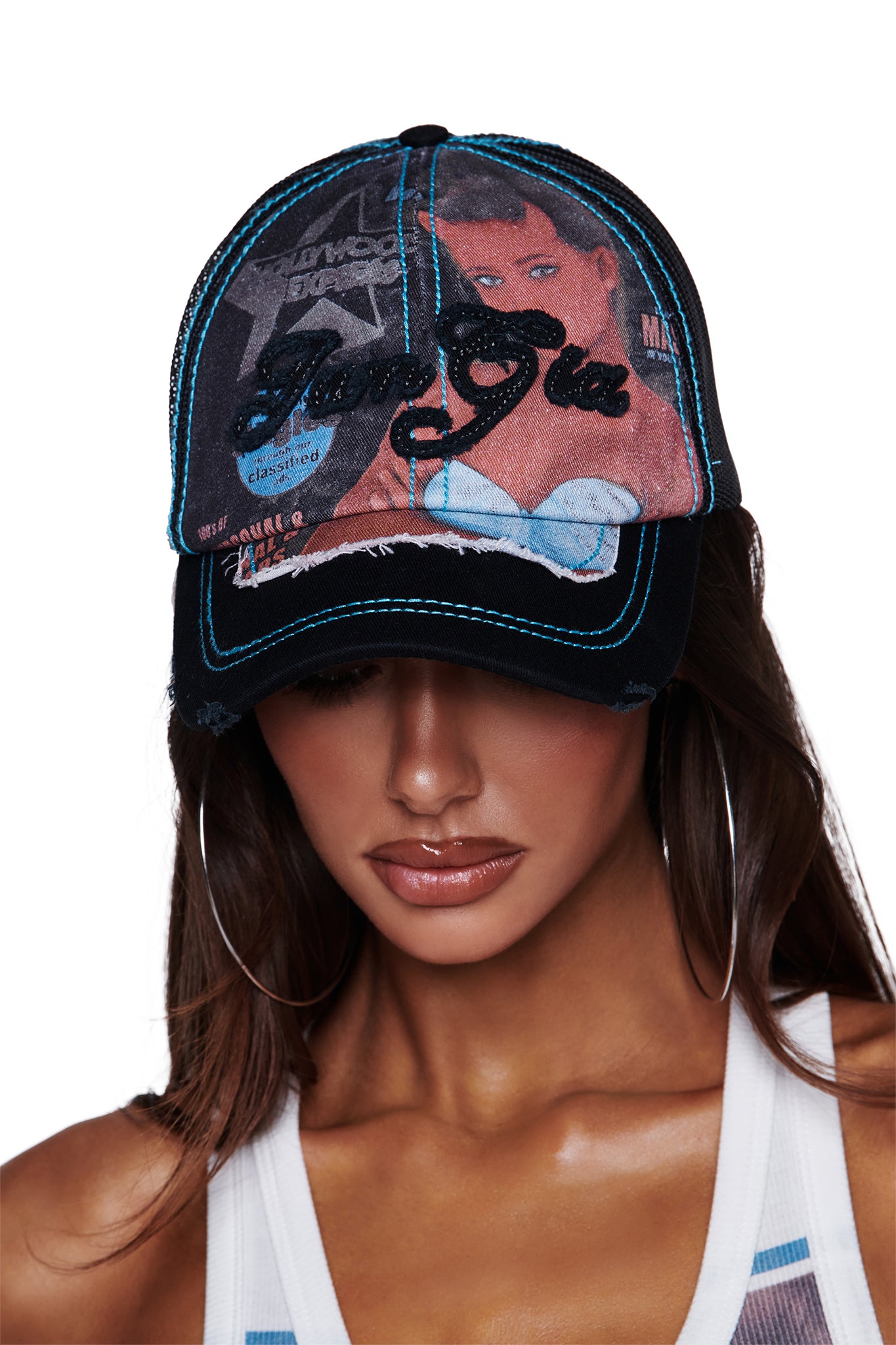 A woman wears the I.AM.GIA T-REX HAT in black, a distressed trucker hat featuring exclusive vintage-inspired artwork of a woman, light blue accents, and bold “GIA GIA” text. Her long brown hair flows down beneath the hat, paired with large hoop earrings and a white tank top. The look is completed with the hat’s adjustable snap closure for a perfect fit.