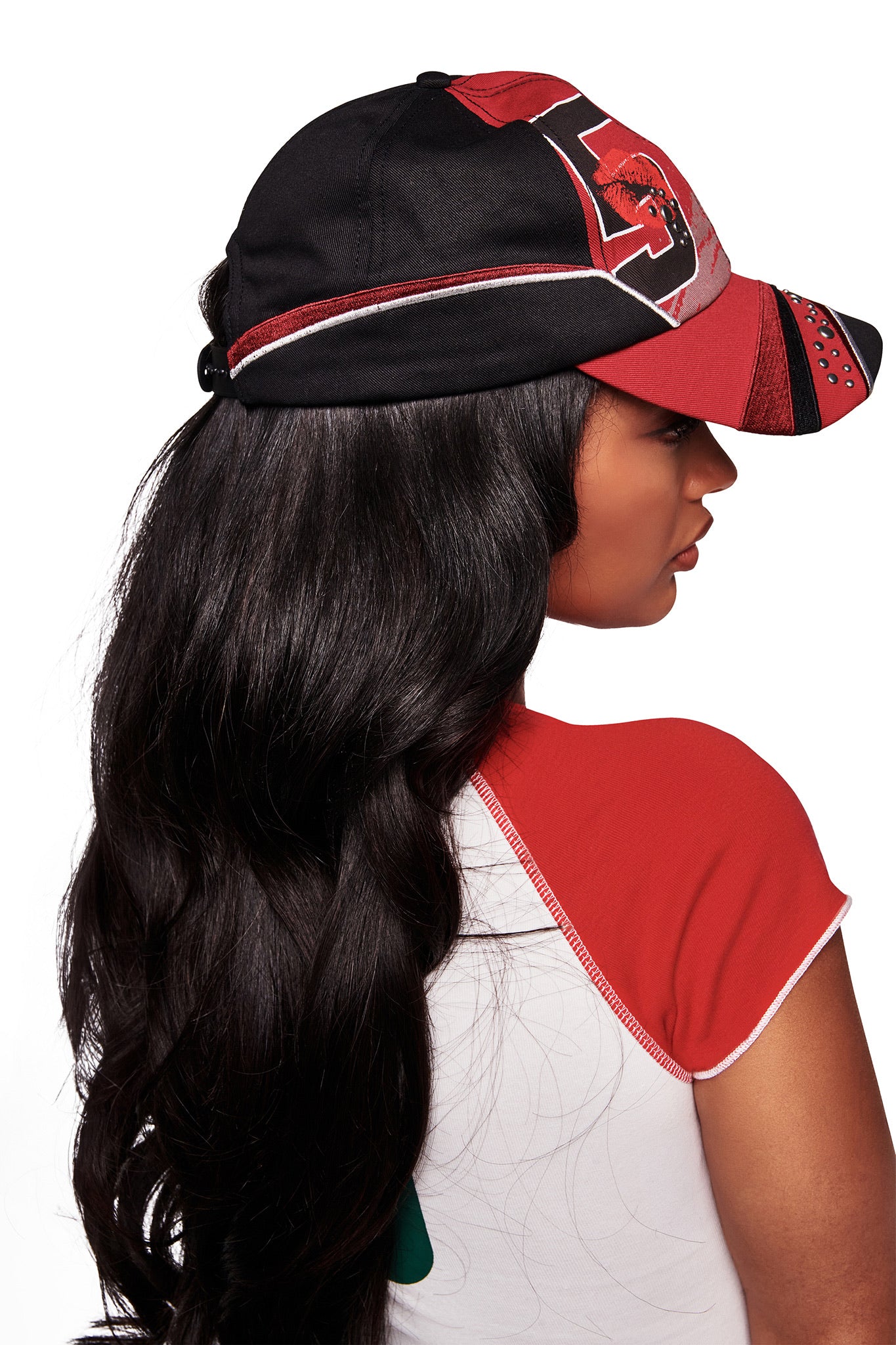 A woman with long, wavy black hair wears the I.AM.GIA SERENA HAT - RED, a black and red racing cap with an adjustable strap back, paired with a white shirt with red sleeves, facing away against a white background.