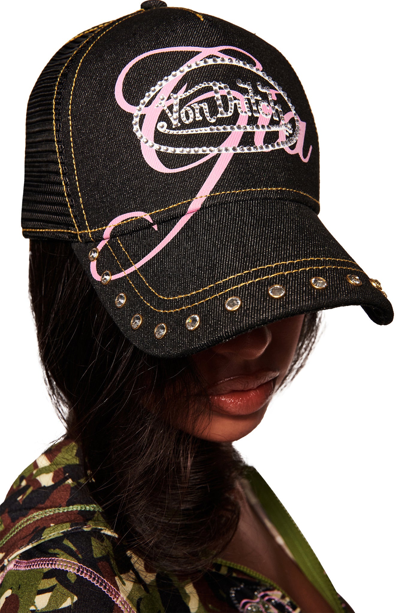 A woman in a camo-pattern top wears the I.AM.GIA X VON DUTCH TRUCKER HAT - INDIGO DENIM, its brim tilted low to partially shade her face.