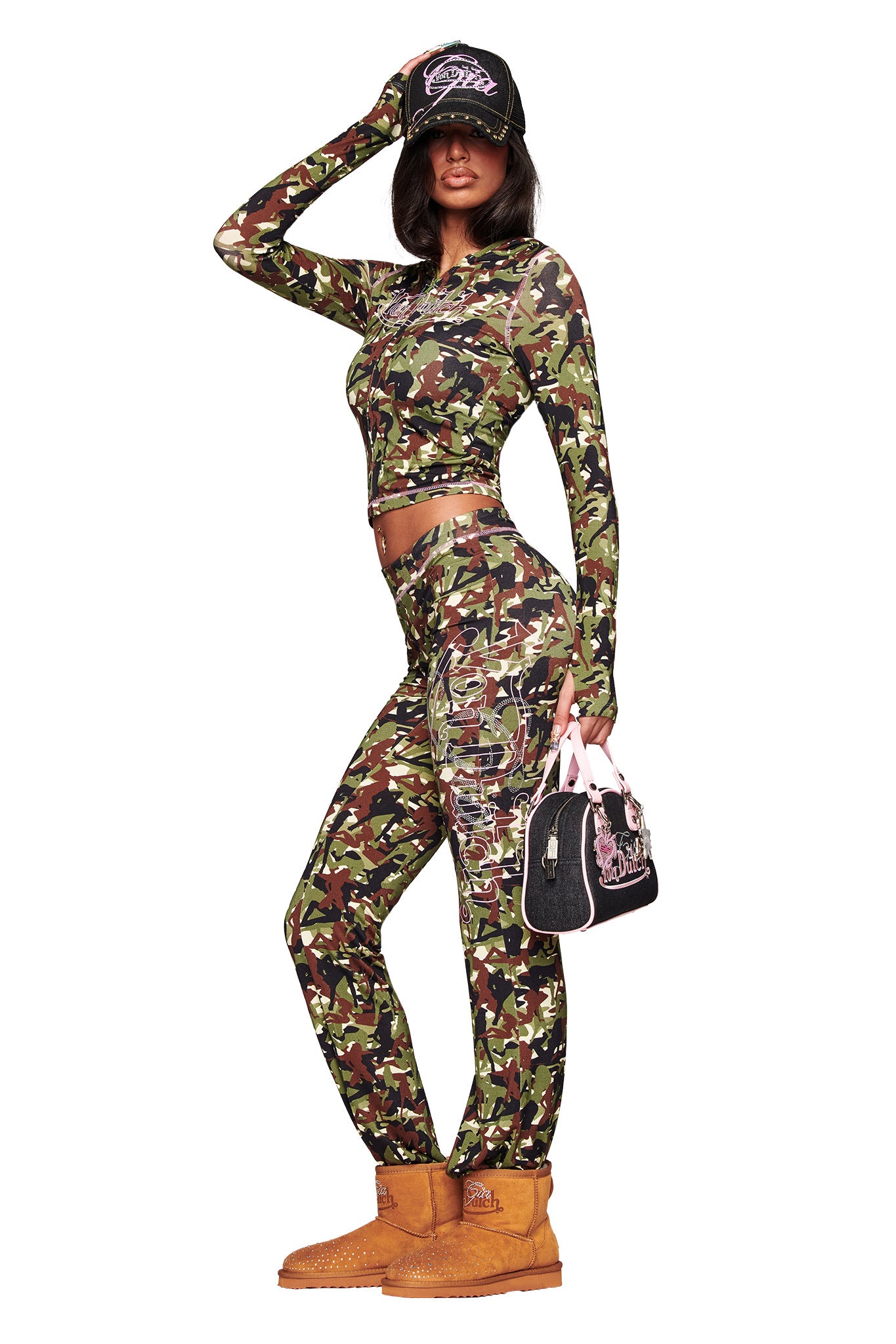 A woman models the I.AM.GIA X VON DUTCH BLARE HOODIE in Gia Girl Camo by I.AM.GIA with matching pants, tan boots, a black cap, sunglasses, and a small black and pink Von Dutch handbag against a white background.