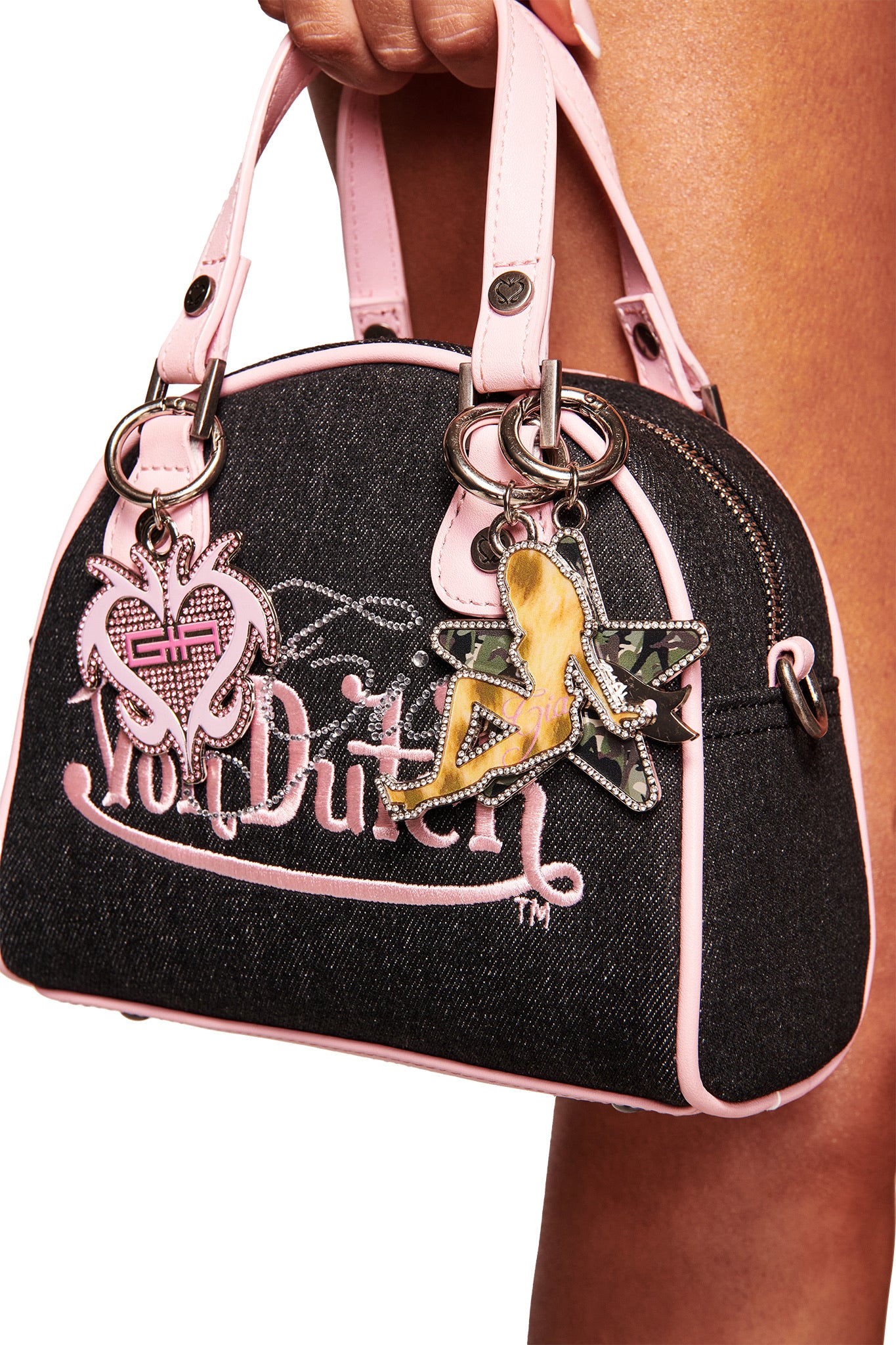 A hand holds a Von Dutch denim handbag with light pink trim, styled as the perfect Y2K accessory. It features two keychains: a rhinestone pink heart and the I.AM.GIA X VON DUTCH GIRL KEYCHAIN in Leopard Pink Girl.