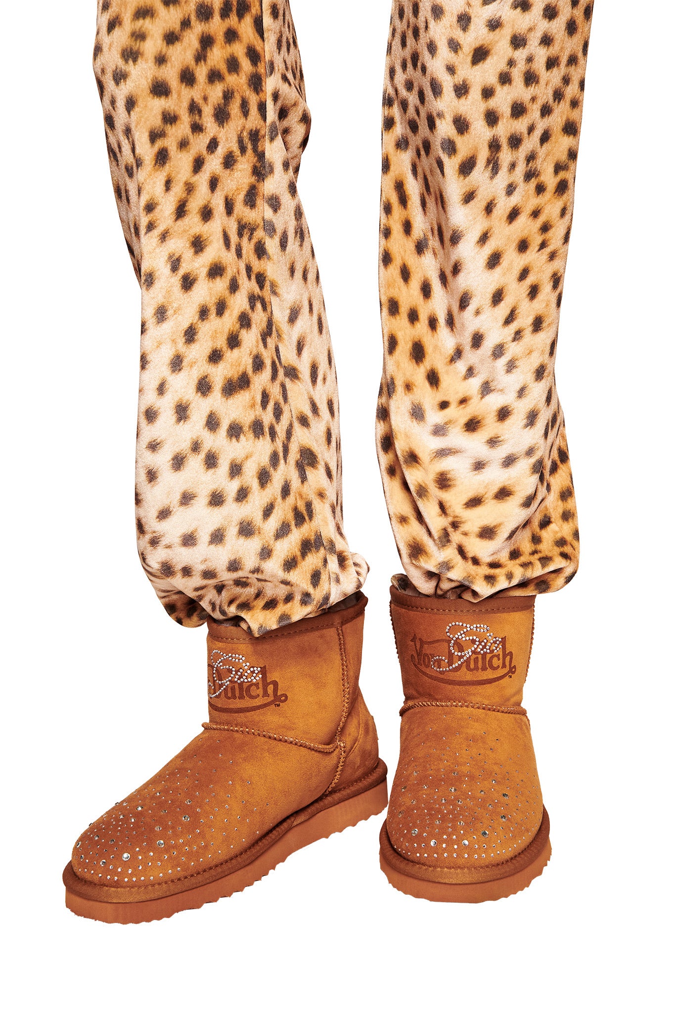 A person wears leopard-print pants and I.AM.GIA X VON DUTCH Faux Shearling Boots in brown, showing only their lower legs and feet. The boots feature rhinestone details and "Koolrich" embroidery on the cuffs.