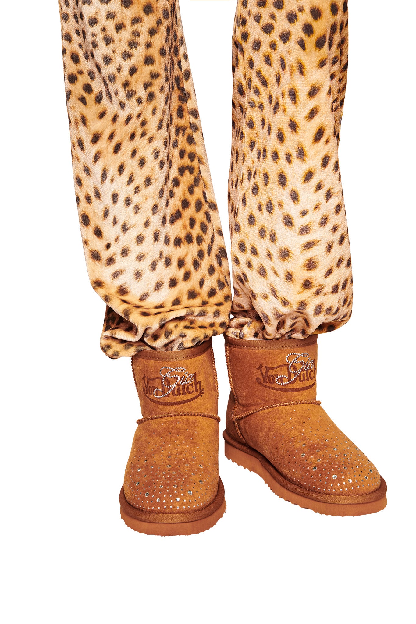 Person in leopard print pants wearing brown I.AM.GIA x Von Dutch Faux Shearling Boot, from the I.AM.GIA collaboration, standing against a white background.