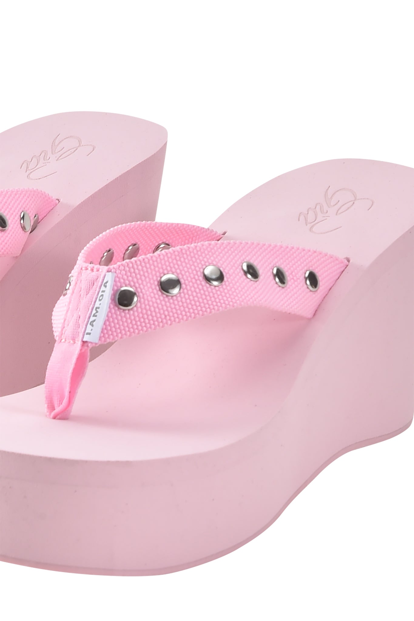 Close-up of the SHELBY STUDDED FLIP FLOP 2.0 - BABY PINK by I.AM.GIA, featuring pink wedge soles, fabric straps with silver studs, and a small white brand tag.