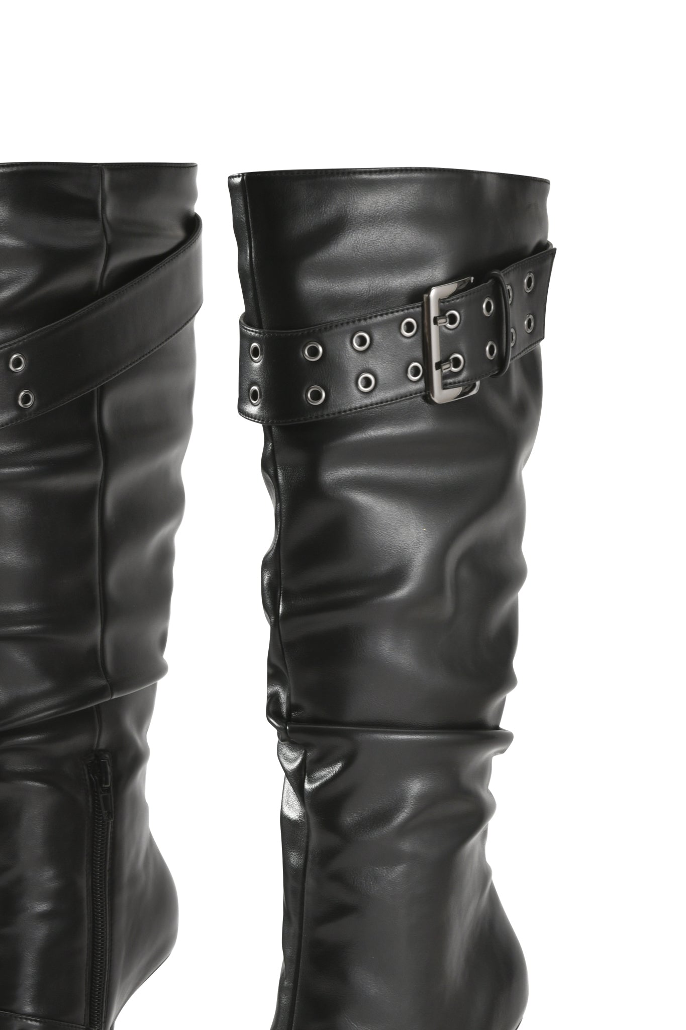 Close-up of the KINTH BOOT 2.0 - BLACK by I.AM.GIA, a slouchy knee-high boot crafted from shiny black faux leather, featuring a decorative strap with silver buckle and grommet details, a black stiletto heel, and visible stitching, shown against a white background.