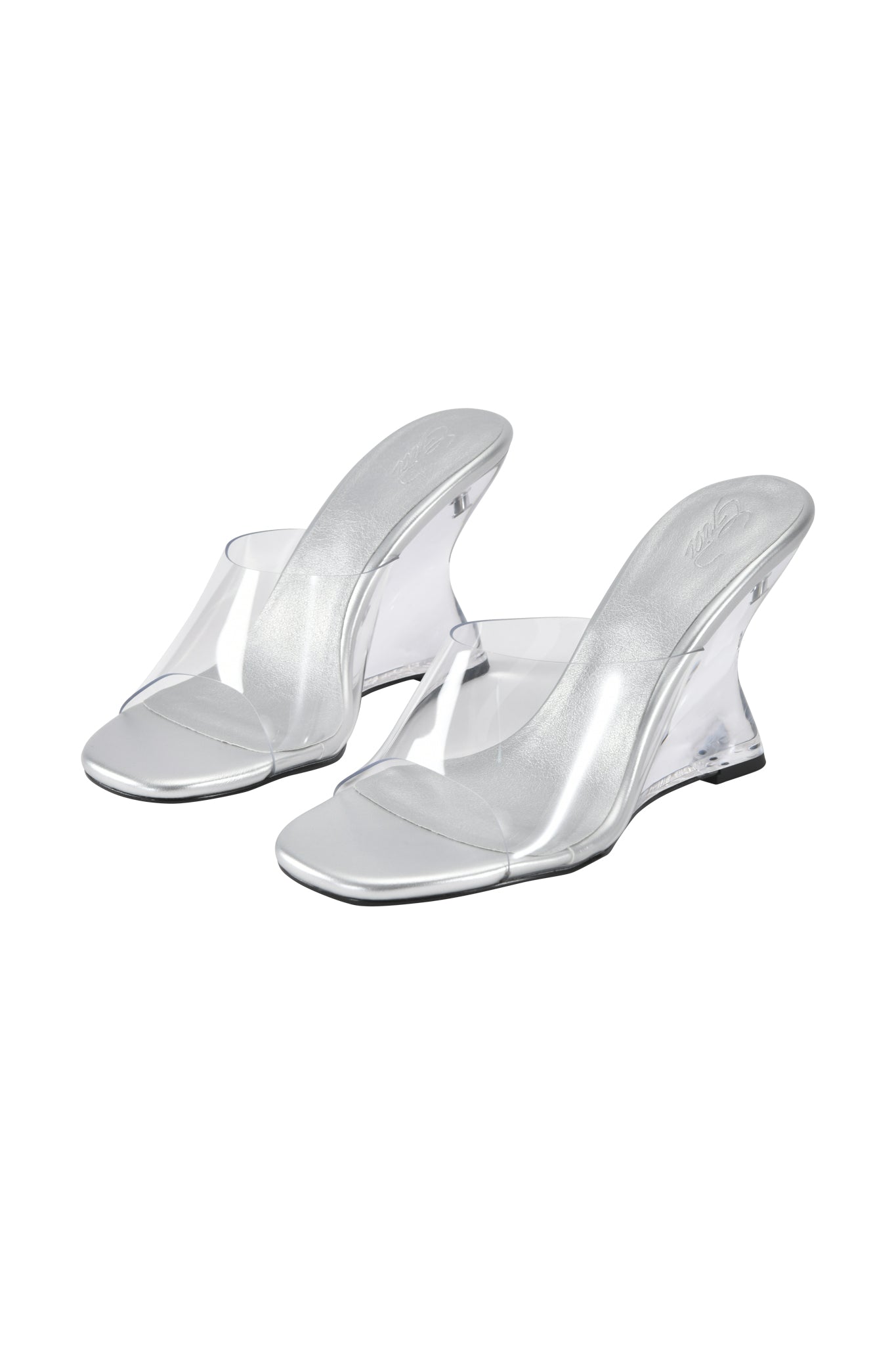The I.AM.GIA RHEA CLEAR WEDGE HEEL features silver sculpted mules with transparent wedge heels and clear vinyl straps, shown against a white background.