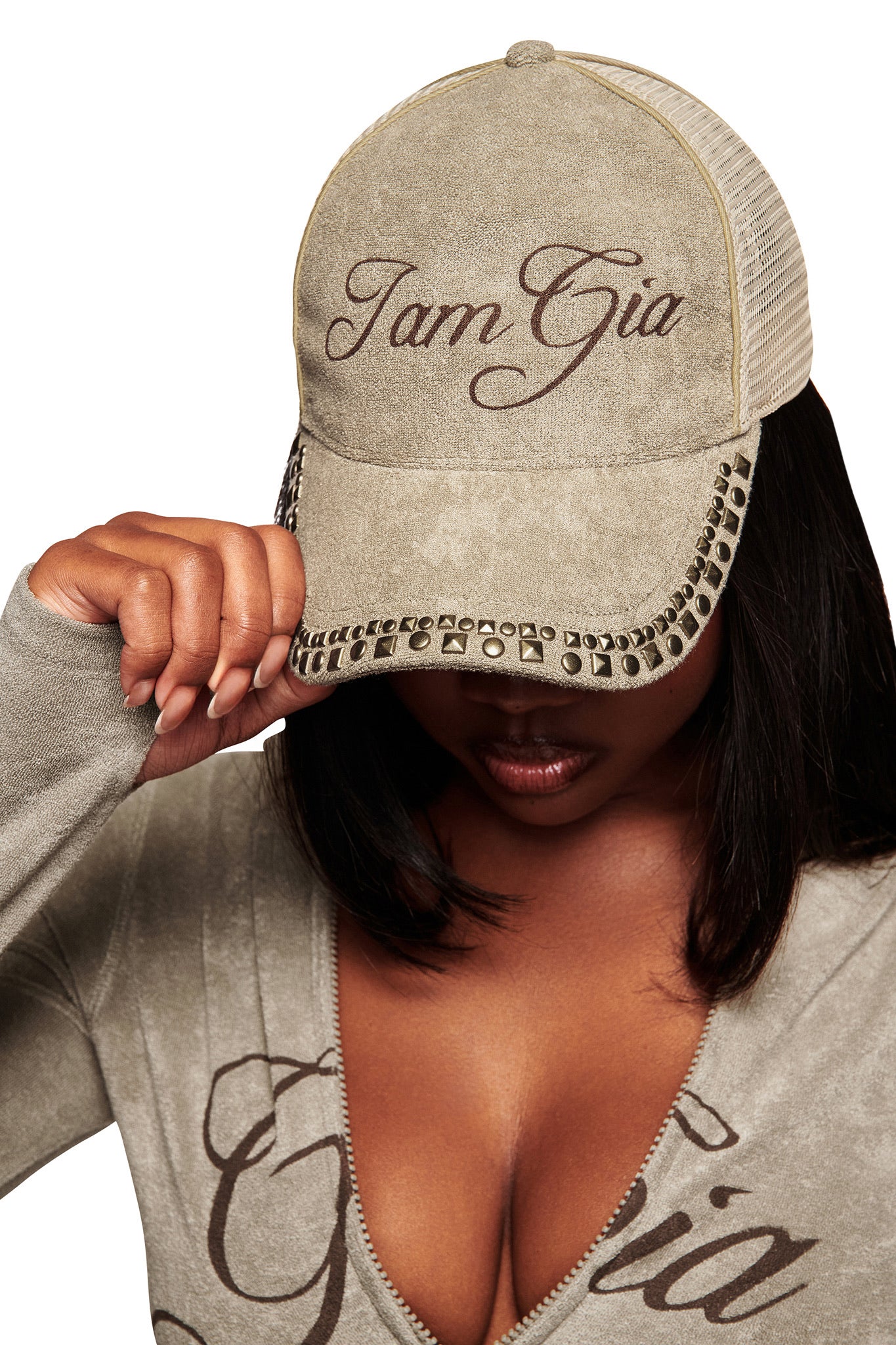 A person wears the ISSHA TERRY HAT - STONE by I.AM.GIA, holding the brim to partially cover their face. They're also dressed in a matching beige deep-neck top with "Gia" printed on it.