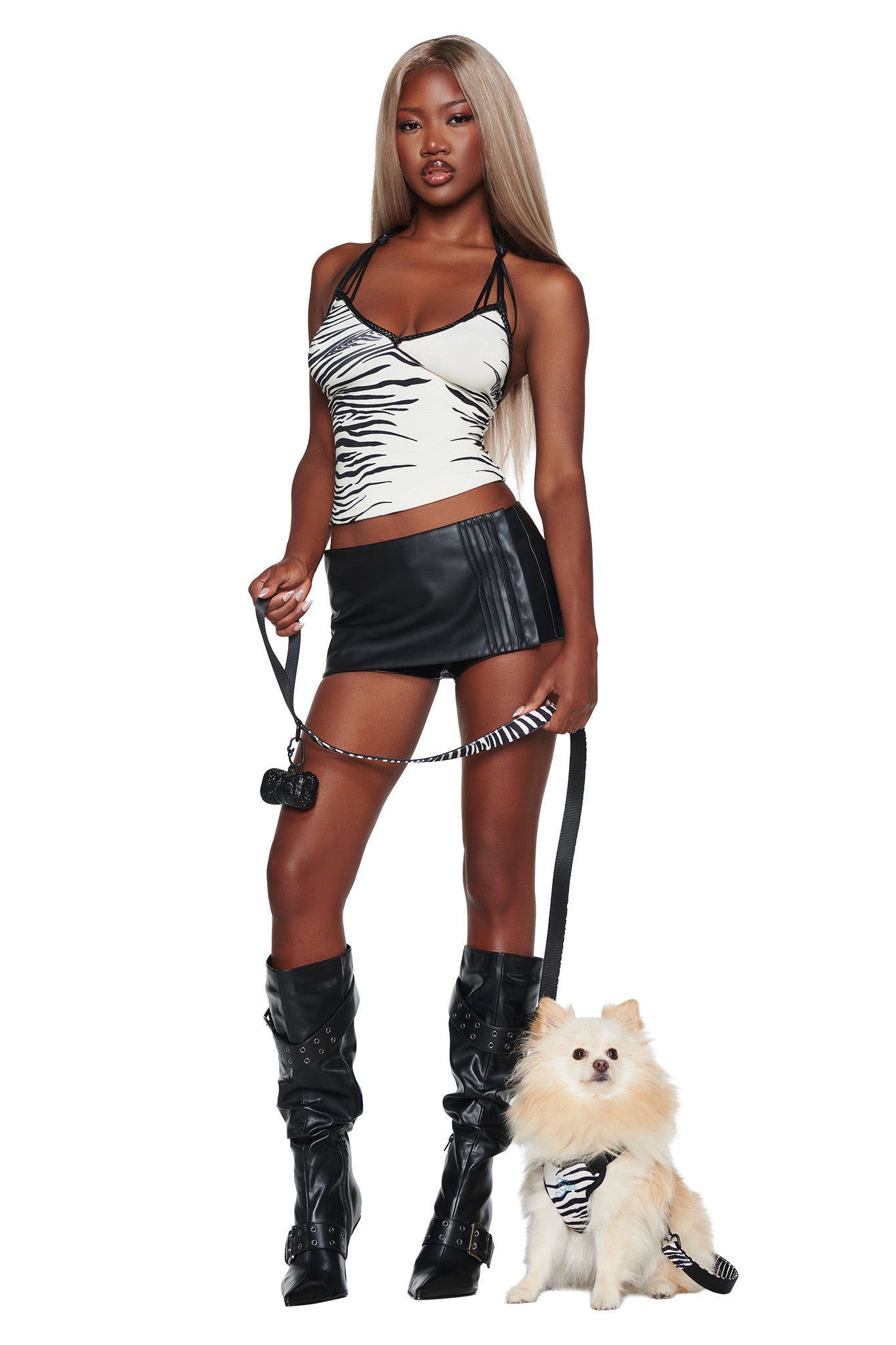 A woman dressed in the I.AM.GIA STAR PAWFECT 3-PIECE SET - FINE LINE ZEBRA stands against a white background, holding the leash of a small white dog outfitted in the matching zebra pet harness.