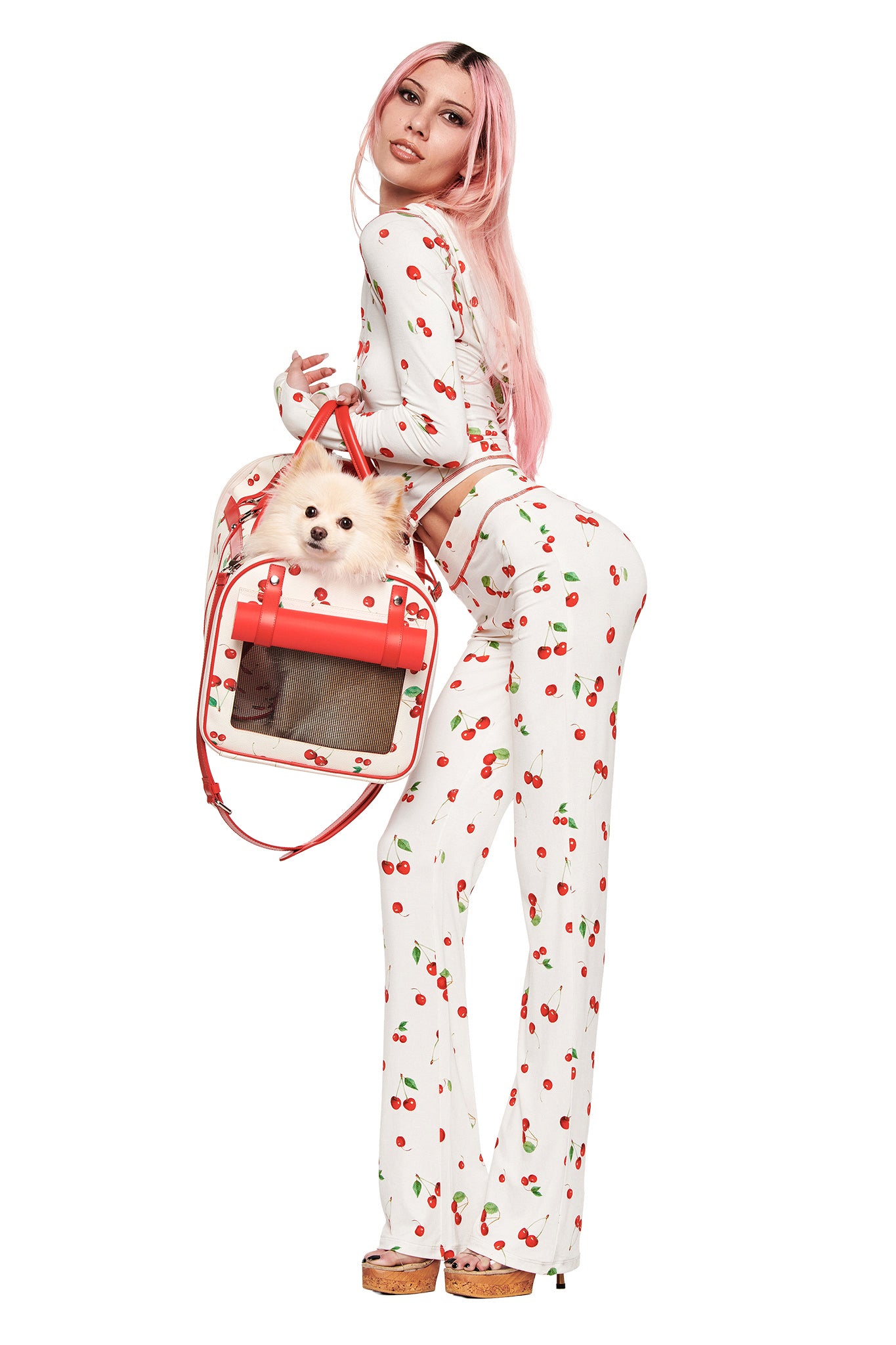 A person with long pink hair, wearing a white cherry print outfit and wooden platform heels, glances over their shoulder while holding a small dog in the I.AM.GIA TRIXX PET CARRIER - CHERRY ON TOP.