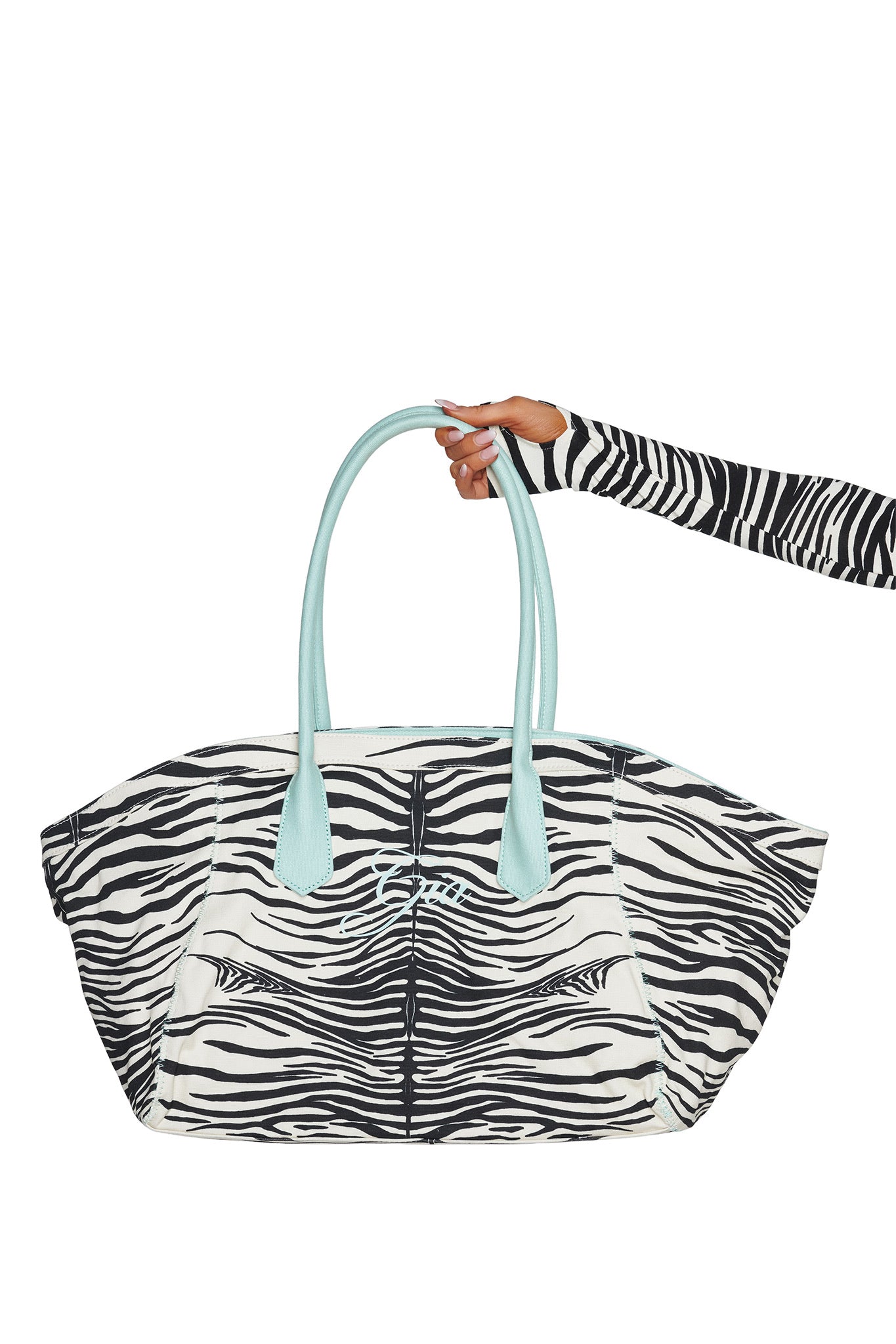 A hand wearing a matching zebra-print sleeve holds the I.AM.GIA BLARE TOTE BAG - FINE LINE ZEBRA, an oversized shopper with light blue handles, against a white background.