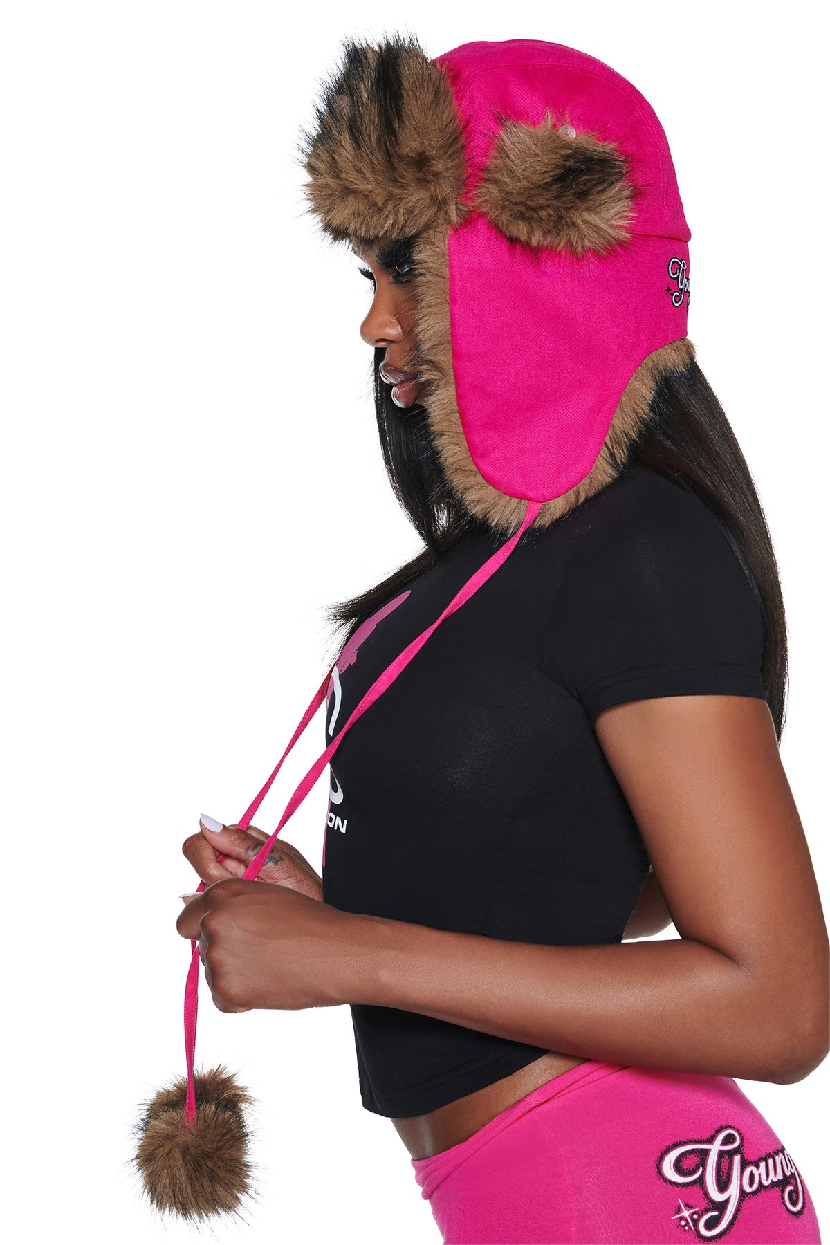 A person wearing an I.AM.GIA YVL FUR TRAPPER HAT - PINK, a bright hot pink trapper hat with brown faux fur trim and dangling pom-poms.