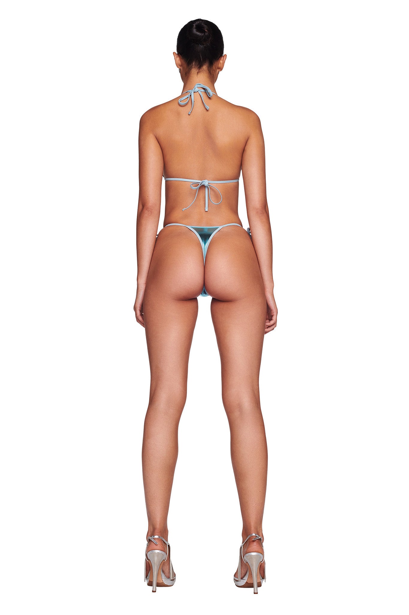 A woman stands with her back to the camera, wearing an I.AM.GIA Alohi Bikini Bottom in blue, with strings tied at the neck and back. She is also sporting silver high heels, and her hair is neatly tied into a bun. The image features a plain white background.