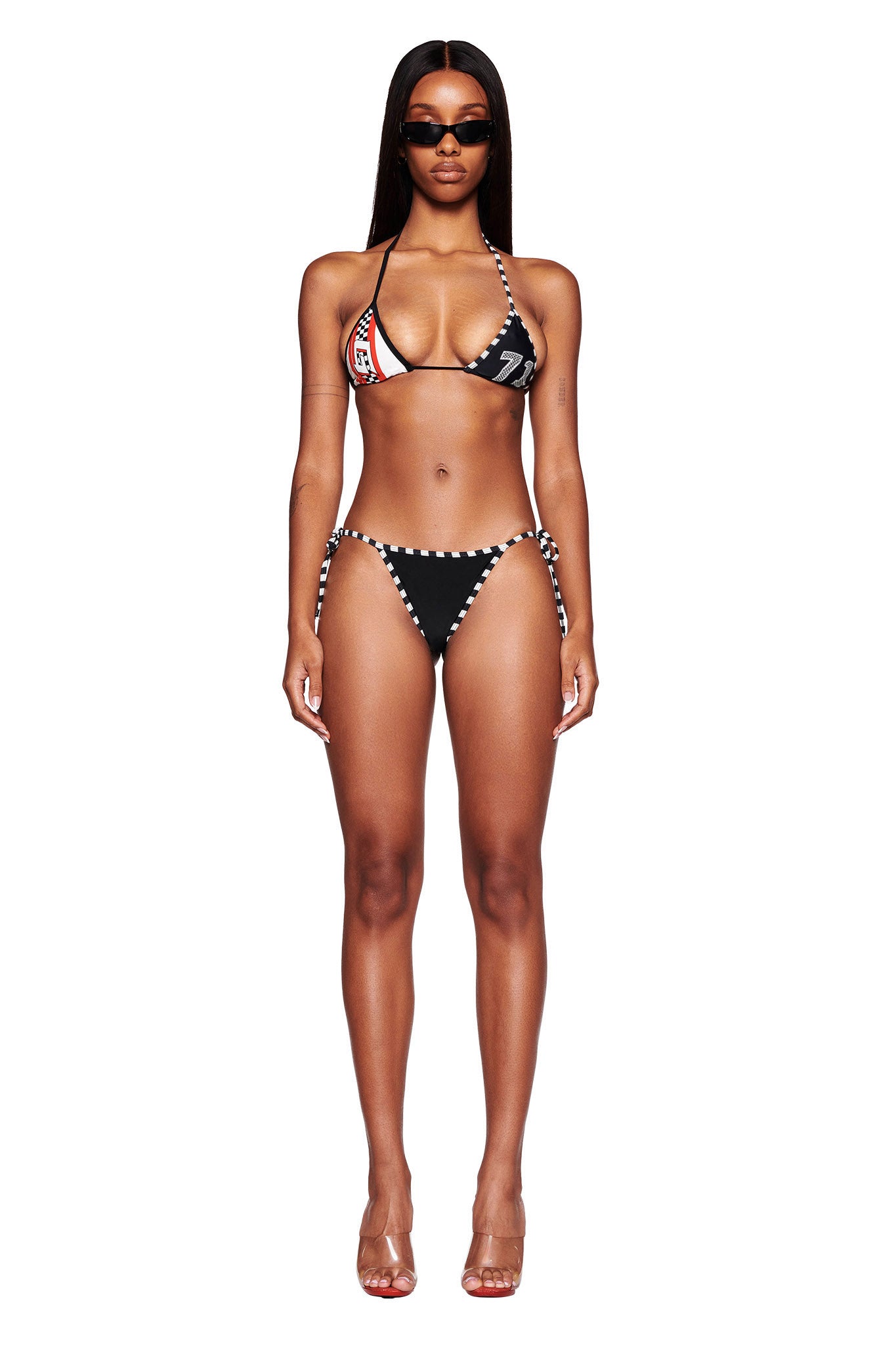 A woman stands facing the camera in I.AM.GIA's RHIANNE BIKINI BOTTOM - BLACK with white accents and a graphic detail. She has long, straight hair and wears dark sunglasses. Complementing her look are clear heeled sandals against a plain white background.