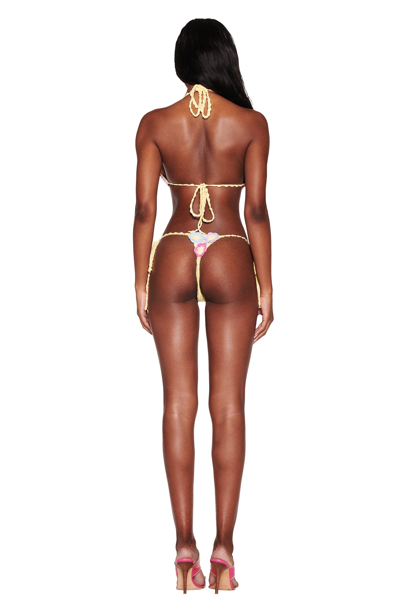 Facing away against a plain white background, a person showcases the intricate design of the BLOSSOM BIKINI TOP - MULTI by I.AM.GIA. The bikini features gold and pink tones with flower bust cups and adjustable ties. Long hair cascades down their back, complementing high heels.